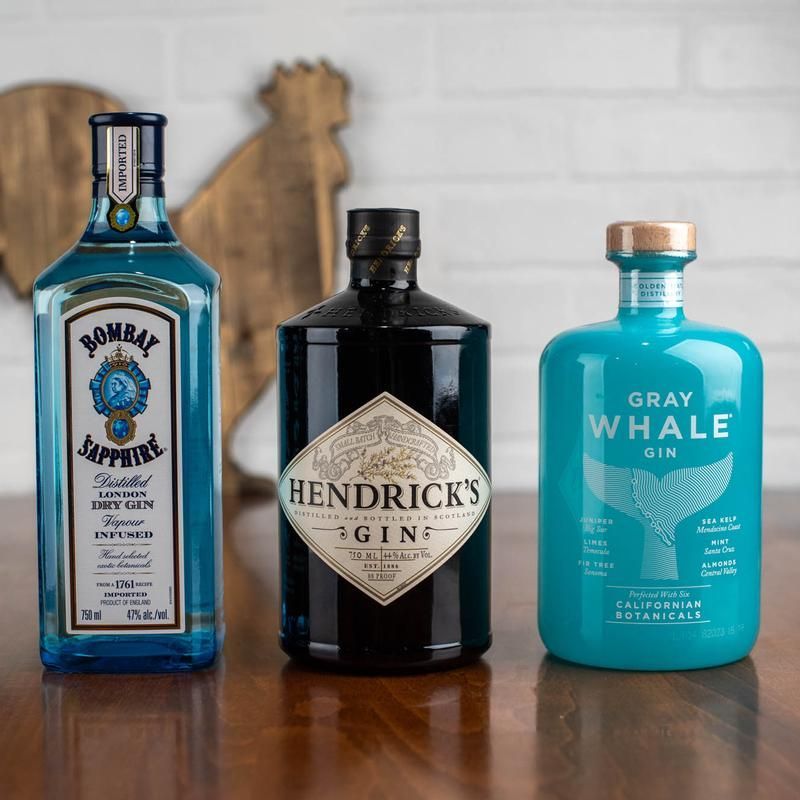 Three gin bottles: Bombay Sapphire, Hendrick's, and Gray Whale on a wooden table with a wooden rooster in the background.