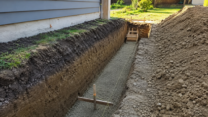 A narrow concrete trench beside a house, bordered by dirt piles and a small chair at the far end.