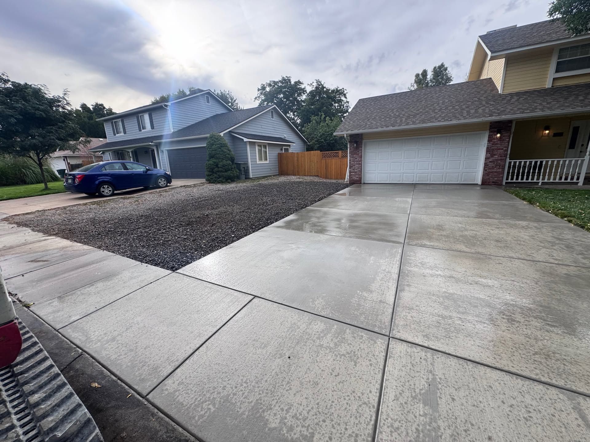 Suburban driveway and two houses, one with a blue car parked nearby, under a cloudy sky