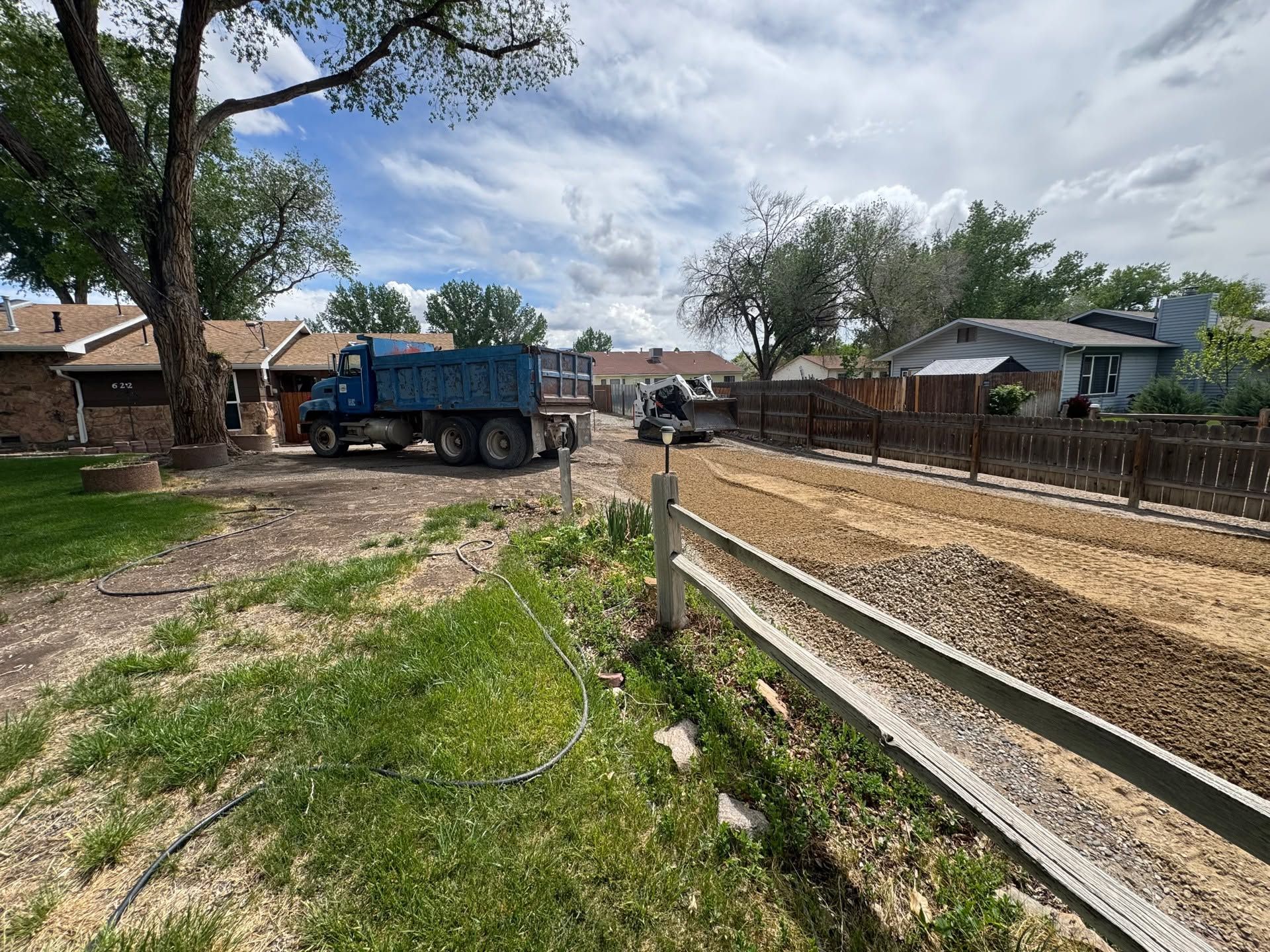 Blue dump truck on a residential dirt road beside a gravel path and wooden fence under a cloudy sky