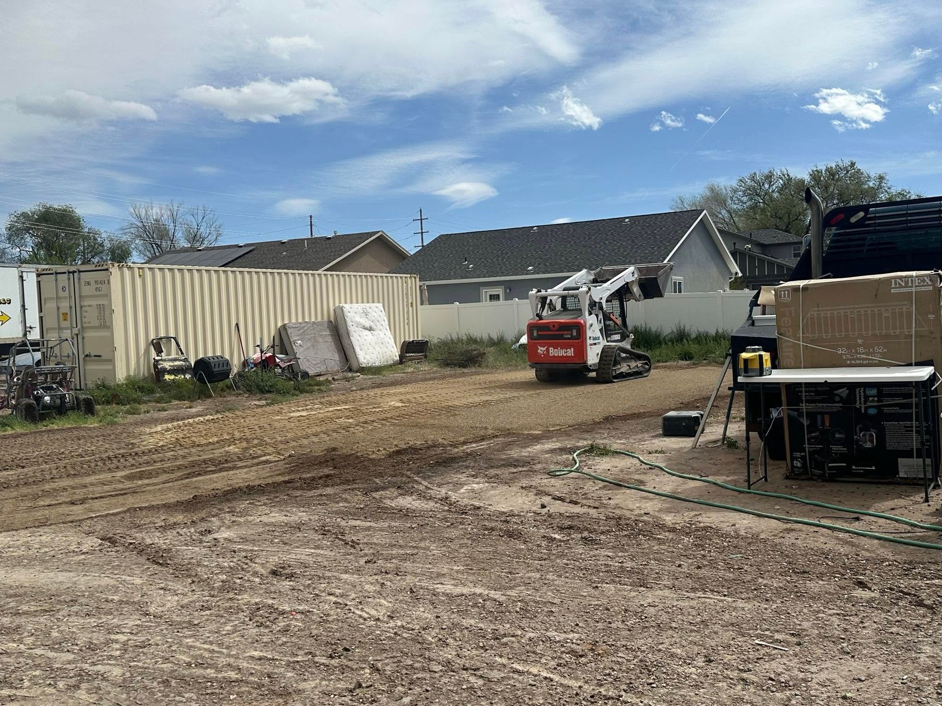 Dirt lot with a red skid steer, a white trailer, and equipment near buildings under a blue sky