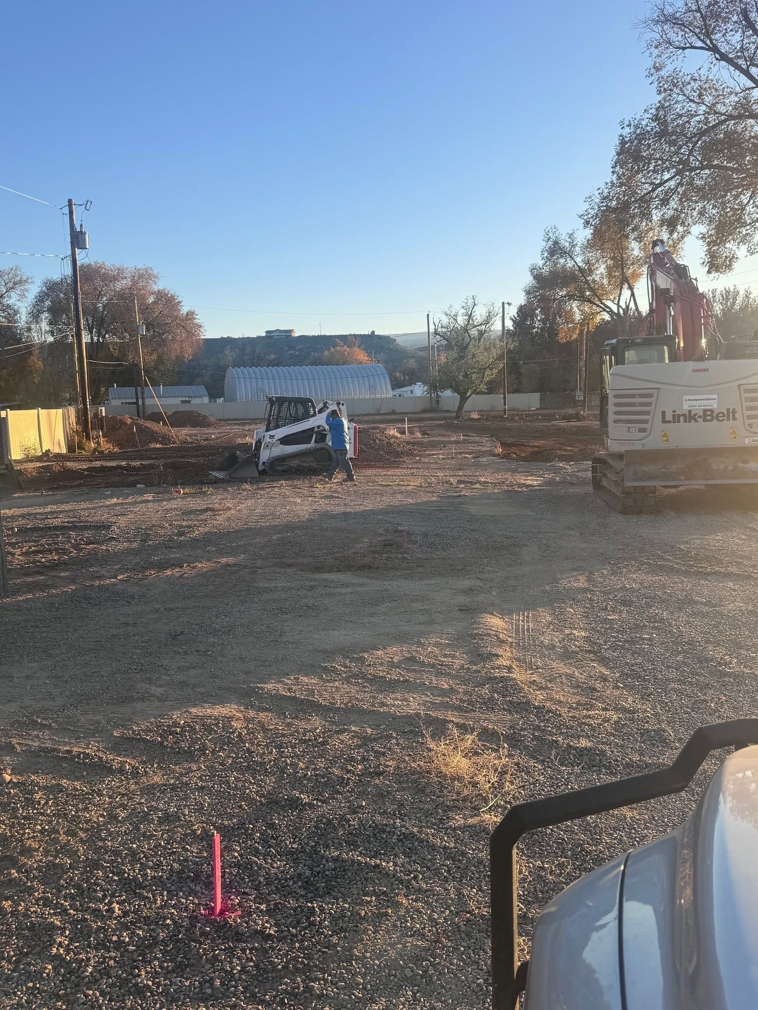 Gravel lot with construction equipment, a white car, and a “Lind…” sign under a clear blue sky.