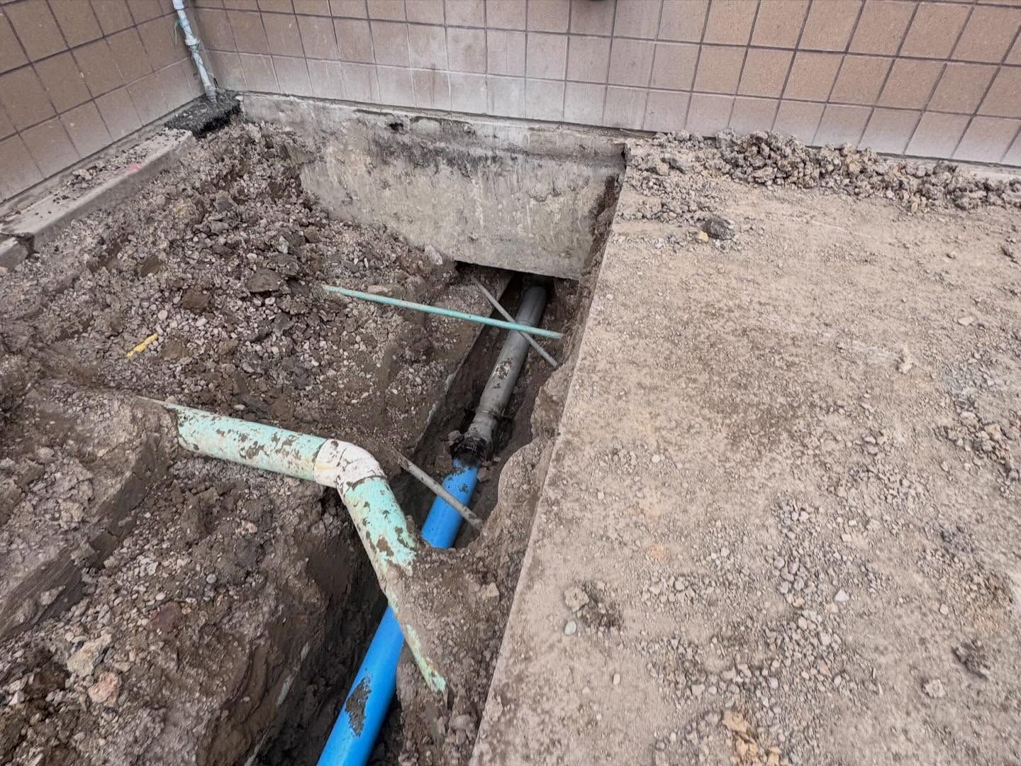 Trench beside tiled wall with exposed pipes and debris, suggesting plumbing repair work