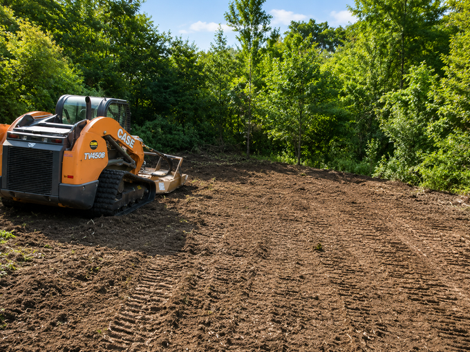 Orange Bobcat skid steer grading a dirt lot beside dense green trees under bright sunlight