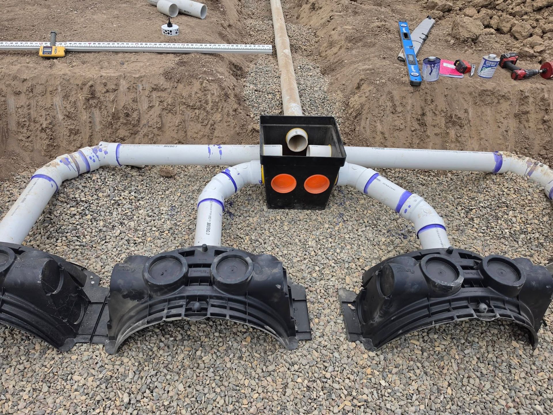 Three black septic tank lids connected by white pipes in a dirt trench with a central junction box.