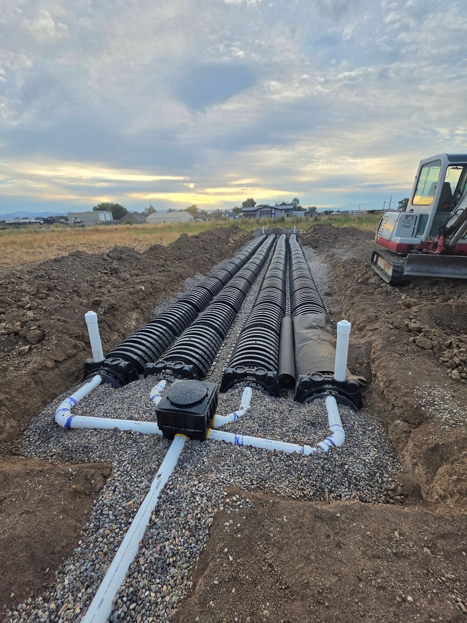 Underground drainage pipes laid in gravel at a construction site, with excavator and open field at sunset
