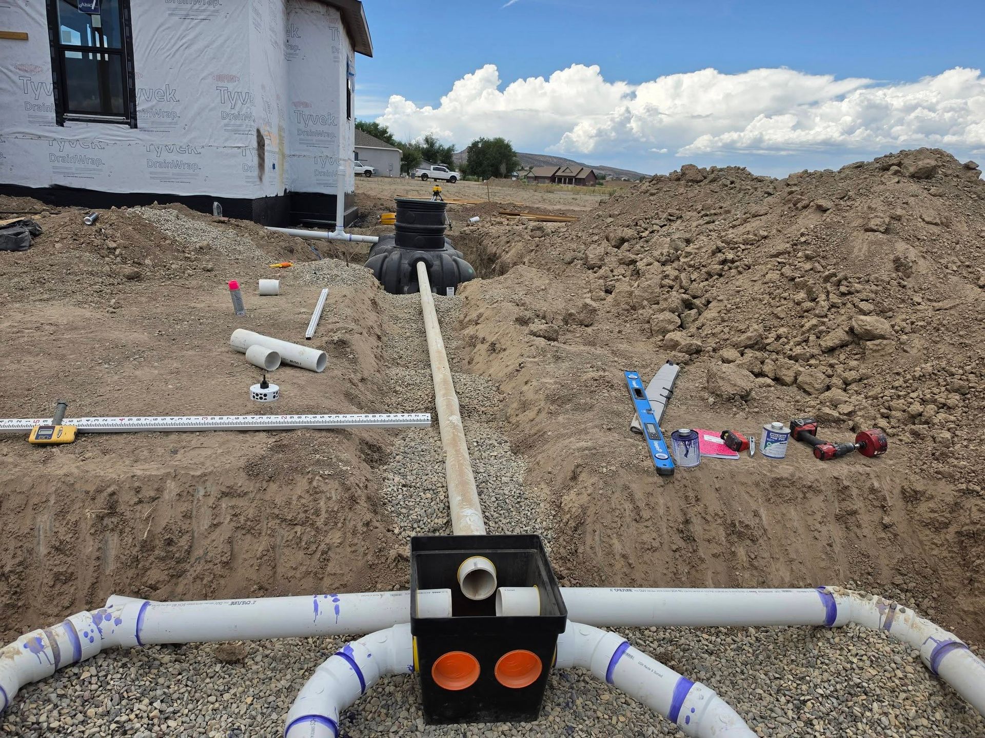 Underground plumbing pipes and fittings laid in a dirt trench beside a house under construction.