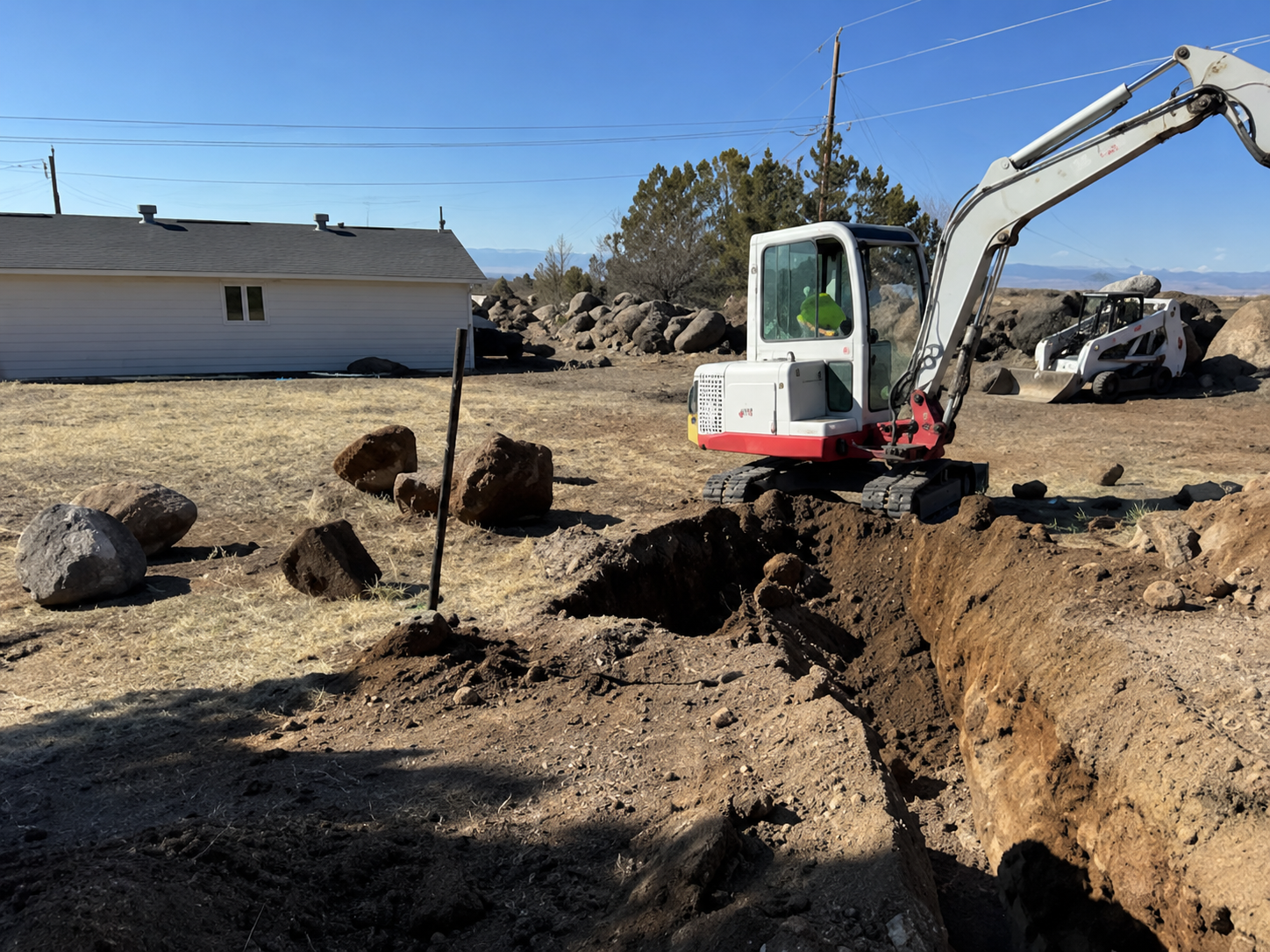 Small excavator digging a trench on a dusty lot beside a white building and piles of dirt