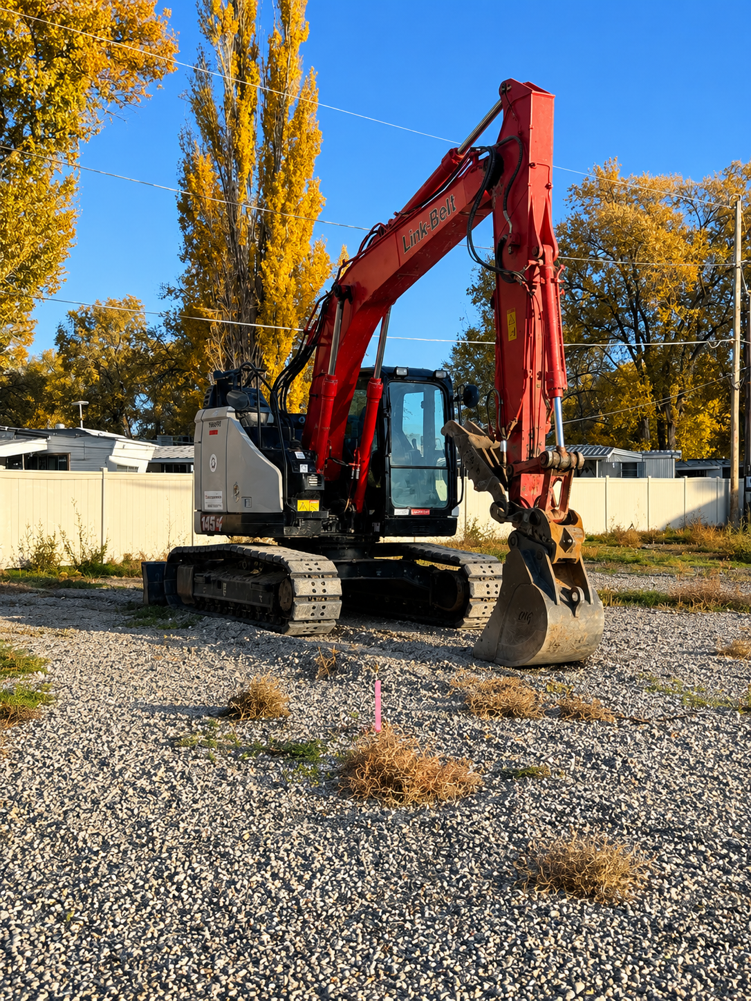 Red excavator on gravel in a yard with autumn trees and a white fence under a clear blue sky