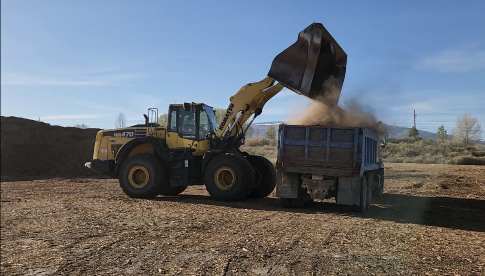 Yellow loader dumping dirt into a truck in a rural field under a blue sky
