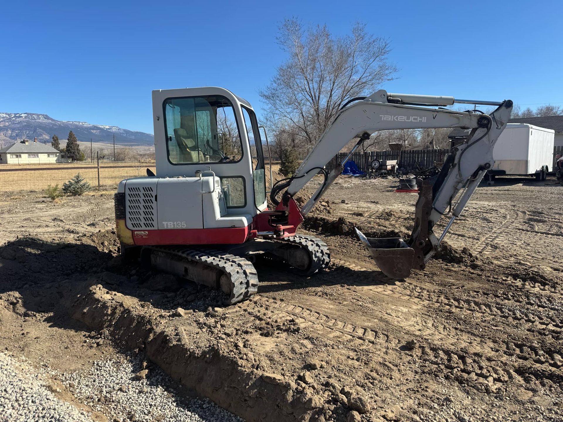Small excavator digging dirt in a field on a clear day, with mountains in the background