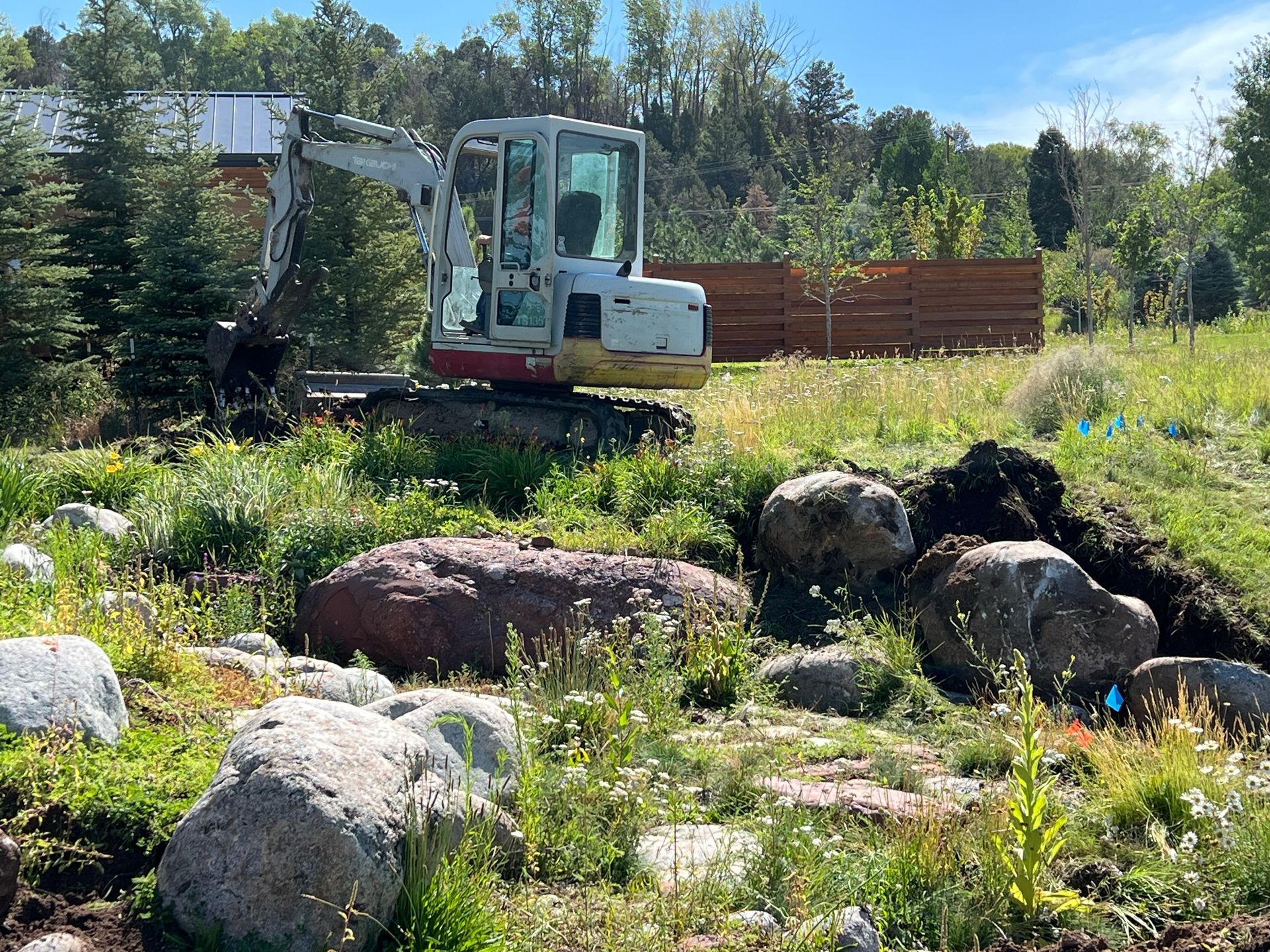 Small excavator by large rocks in a sunny grassy yard with trees and a red fence in the background