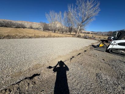 Gravel road being built beside a leafless tree, with a skid steer at right and a shadow in foreground.