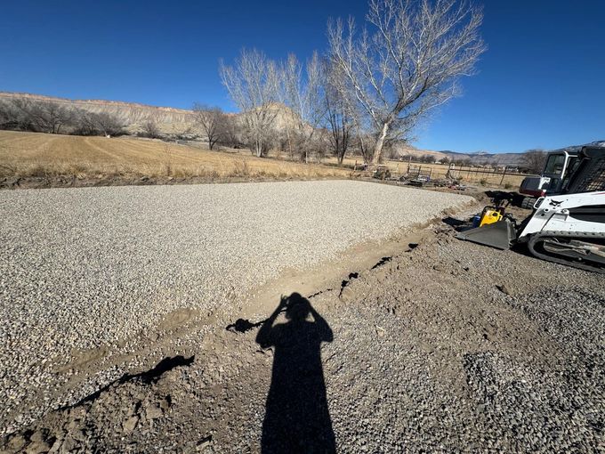 Gravel road construction site with a skid steer, blue sky, and a person’s shadow in the foreground