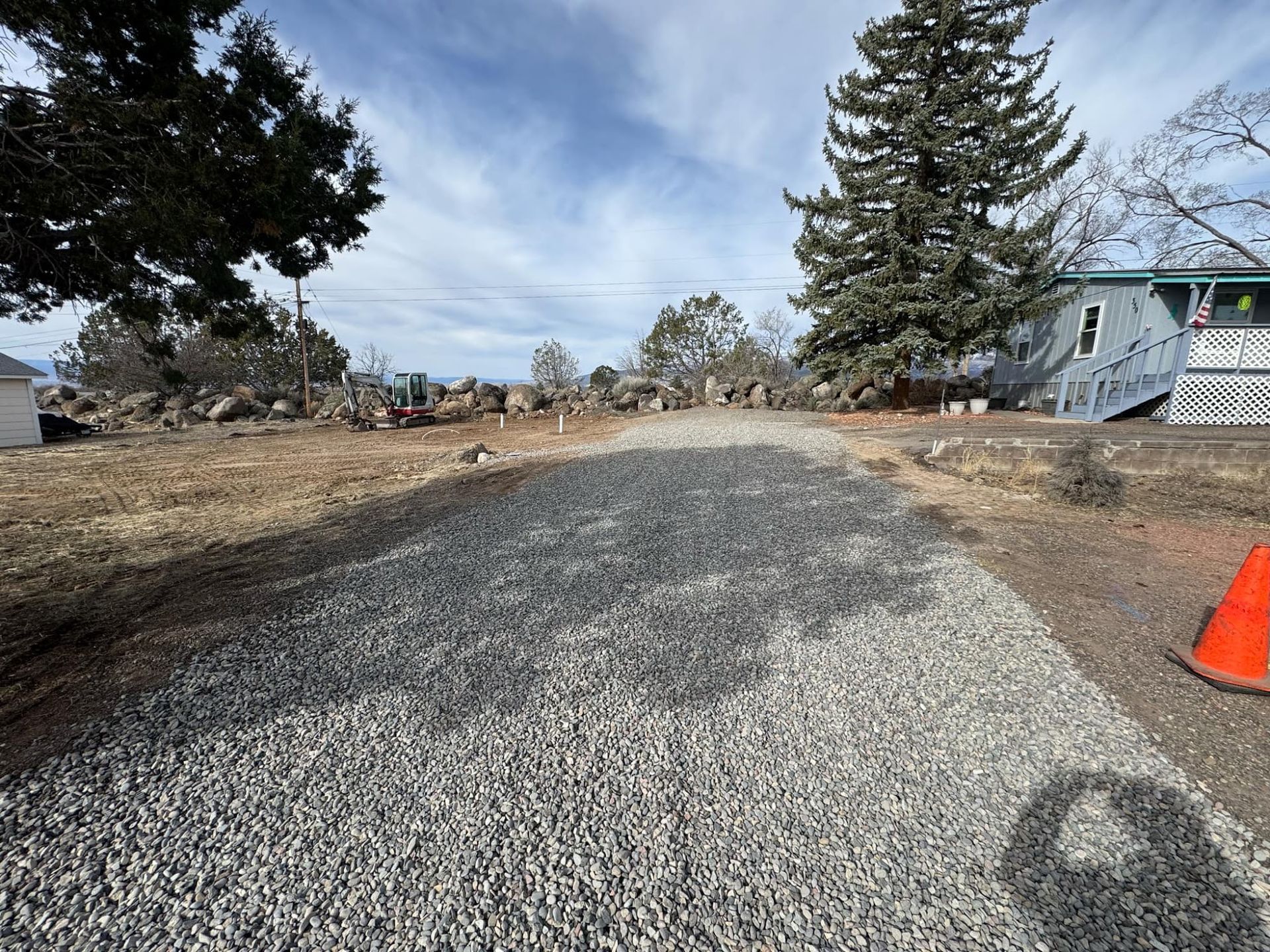 Gravel road under construction with patchy asphalt, trees, houses, and an orange traffic cone on the right.
