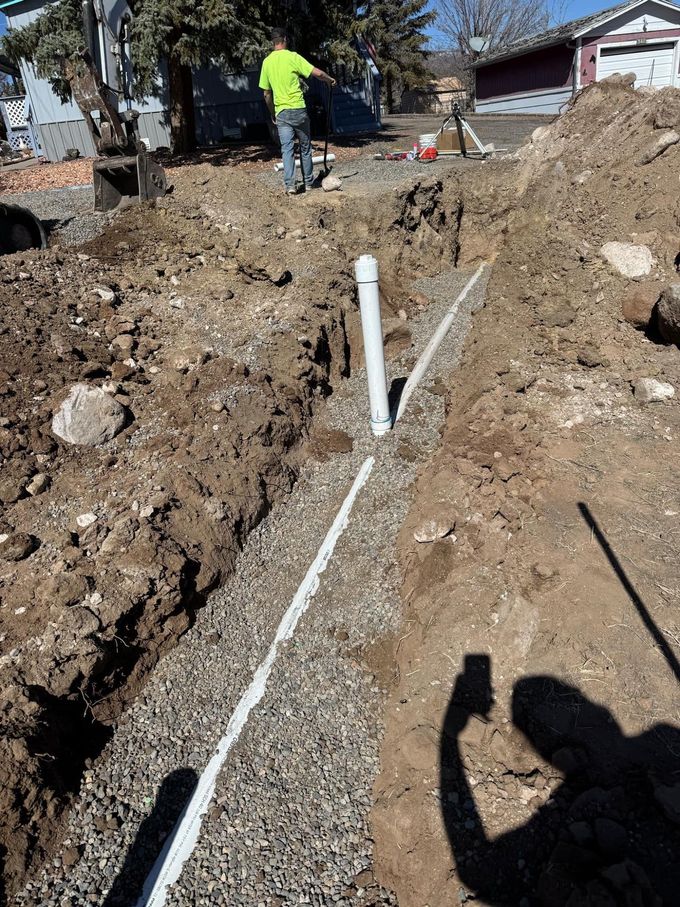 Trench with white PVC pipe and gravel in a yard, with a worker standing in the background.