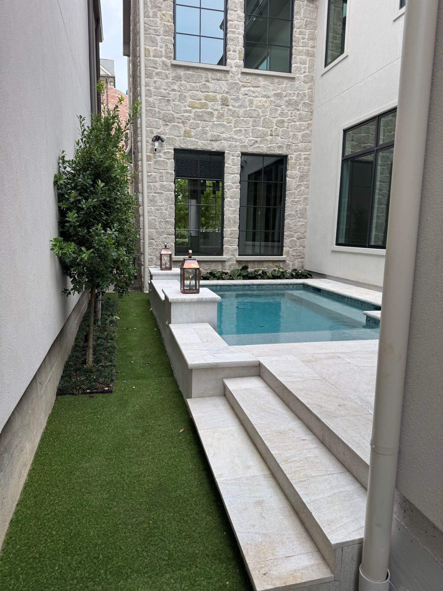 Narrow modern courtyard with a small pool, stone walls, white steps, and a strip of artificial grass