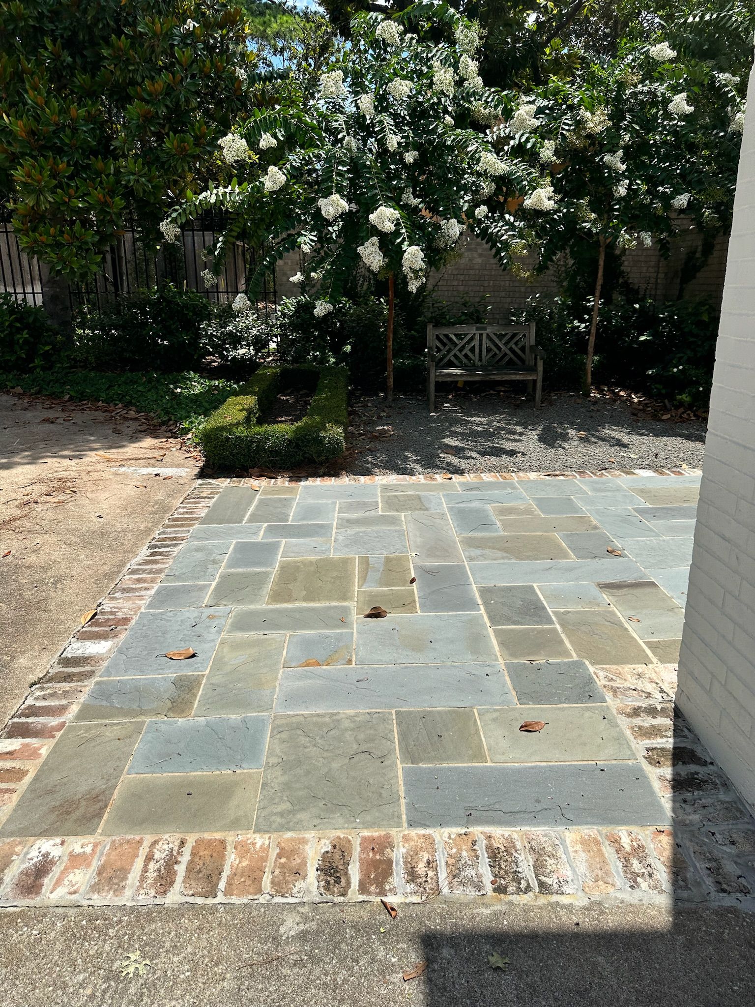Stone patio with a small central marker, bordered by brick, in a garden with trees and a bench