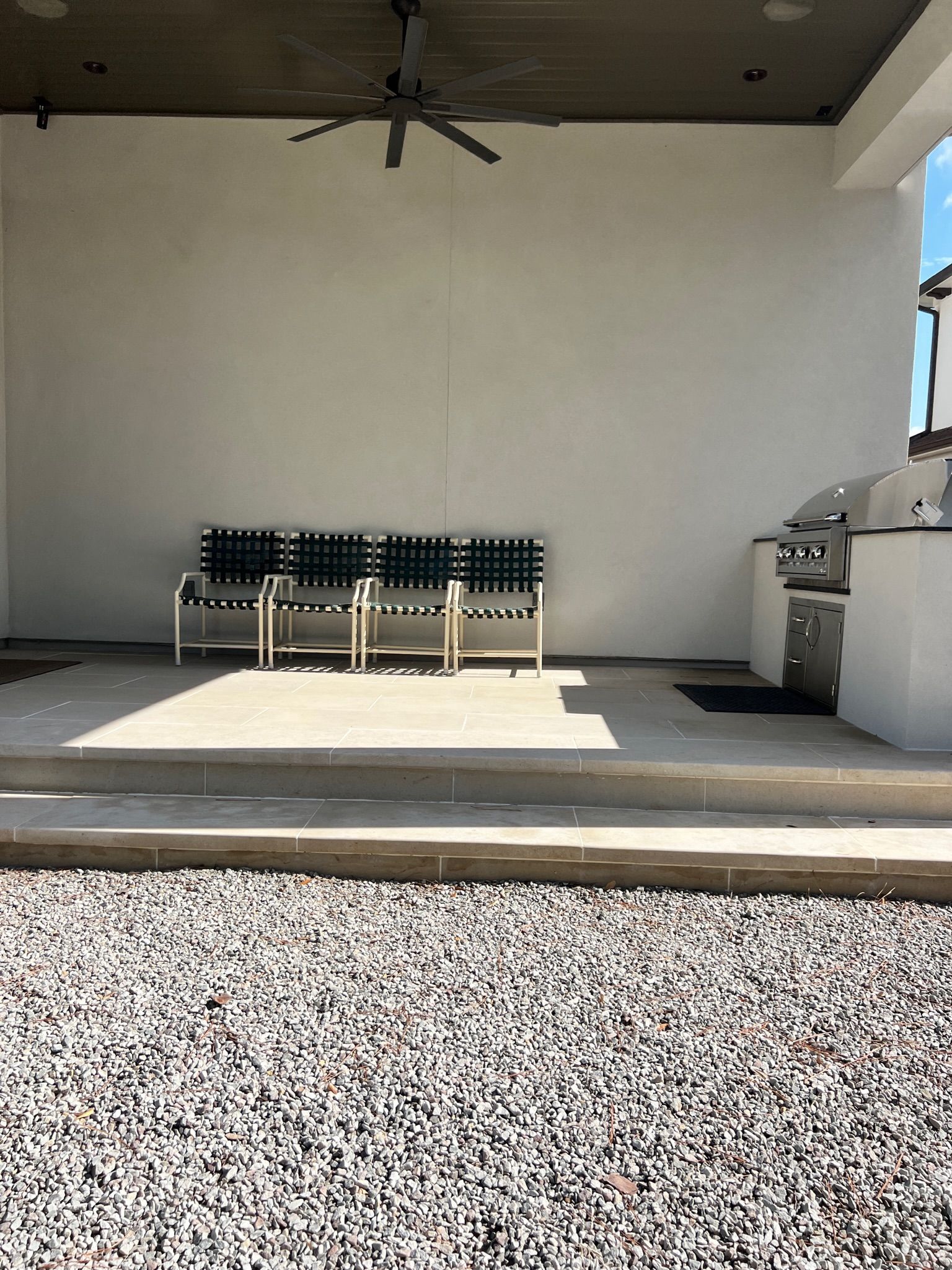 Covered outdoor patio with a ceiling fan, bench seating, and a grill beside a gravel path.