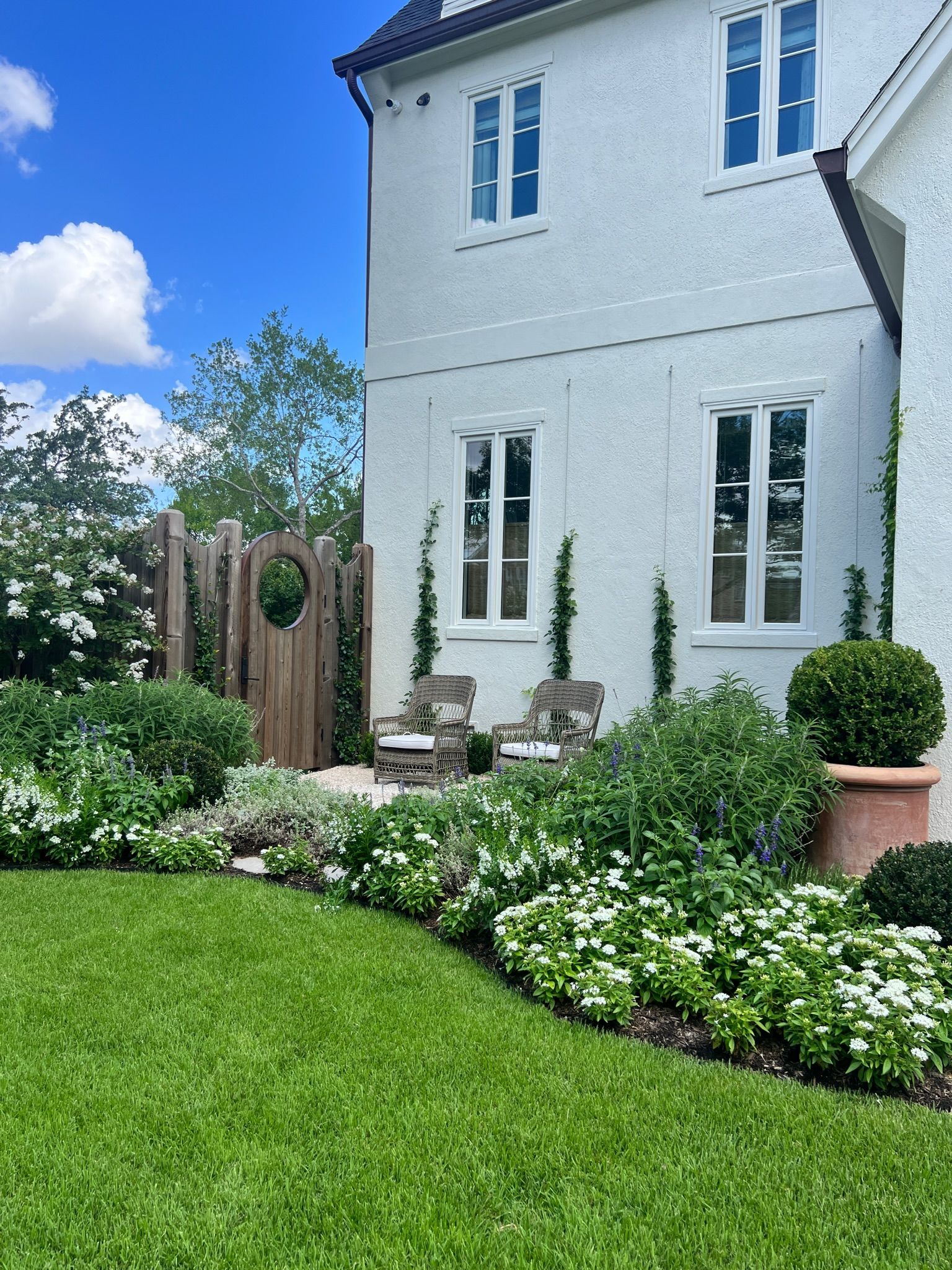 White house with blue trim and manicured flower beds beside a green lawn under a sunny sky