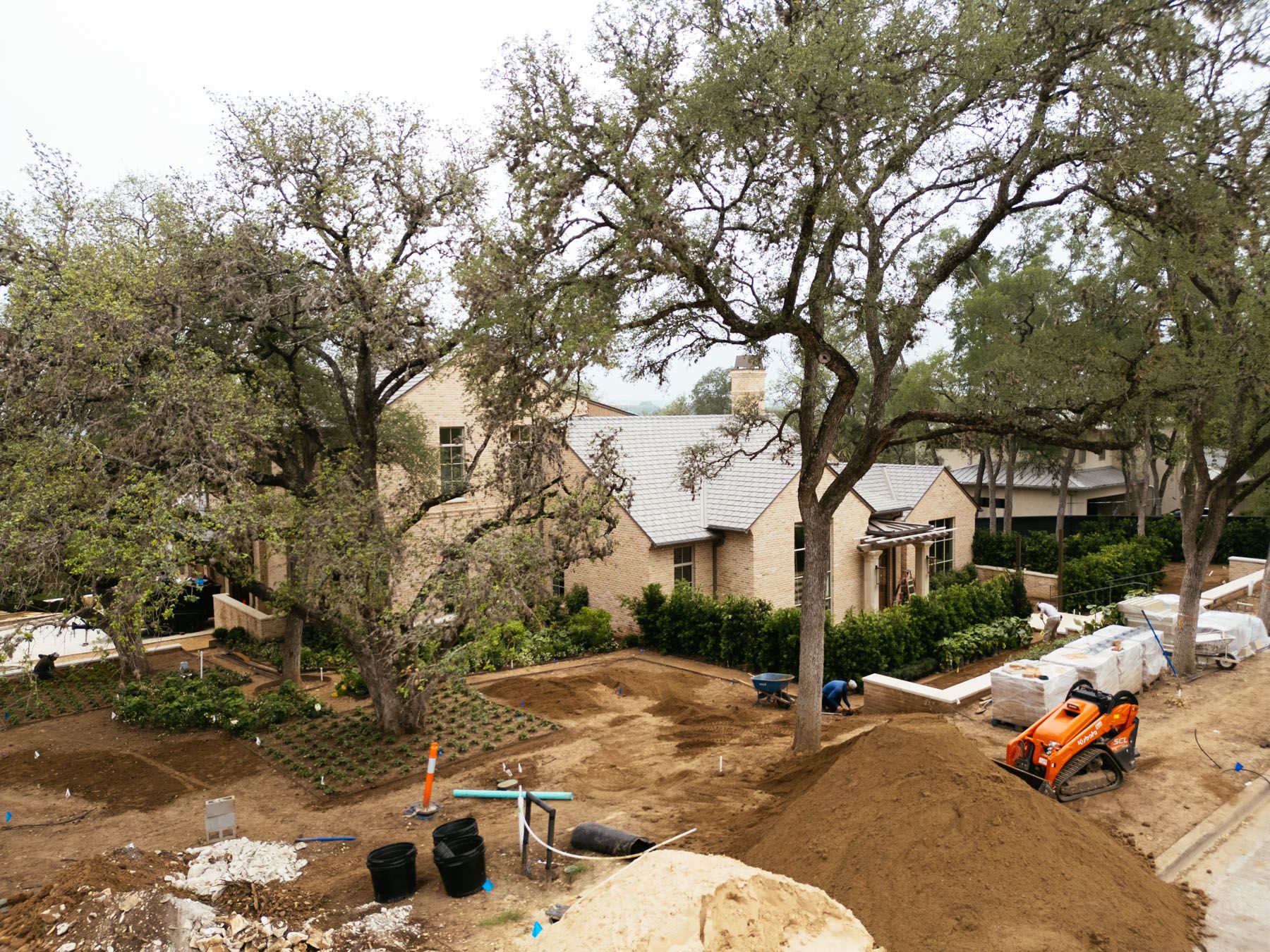 Residential construction site with dirt piles, trees, and an orange excavator beside a house
