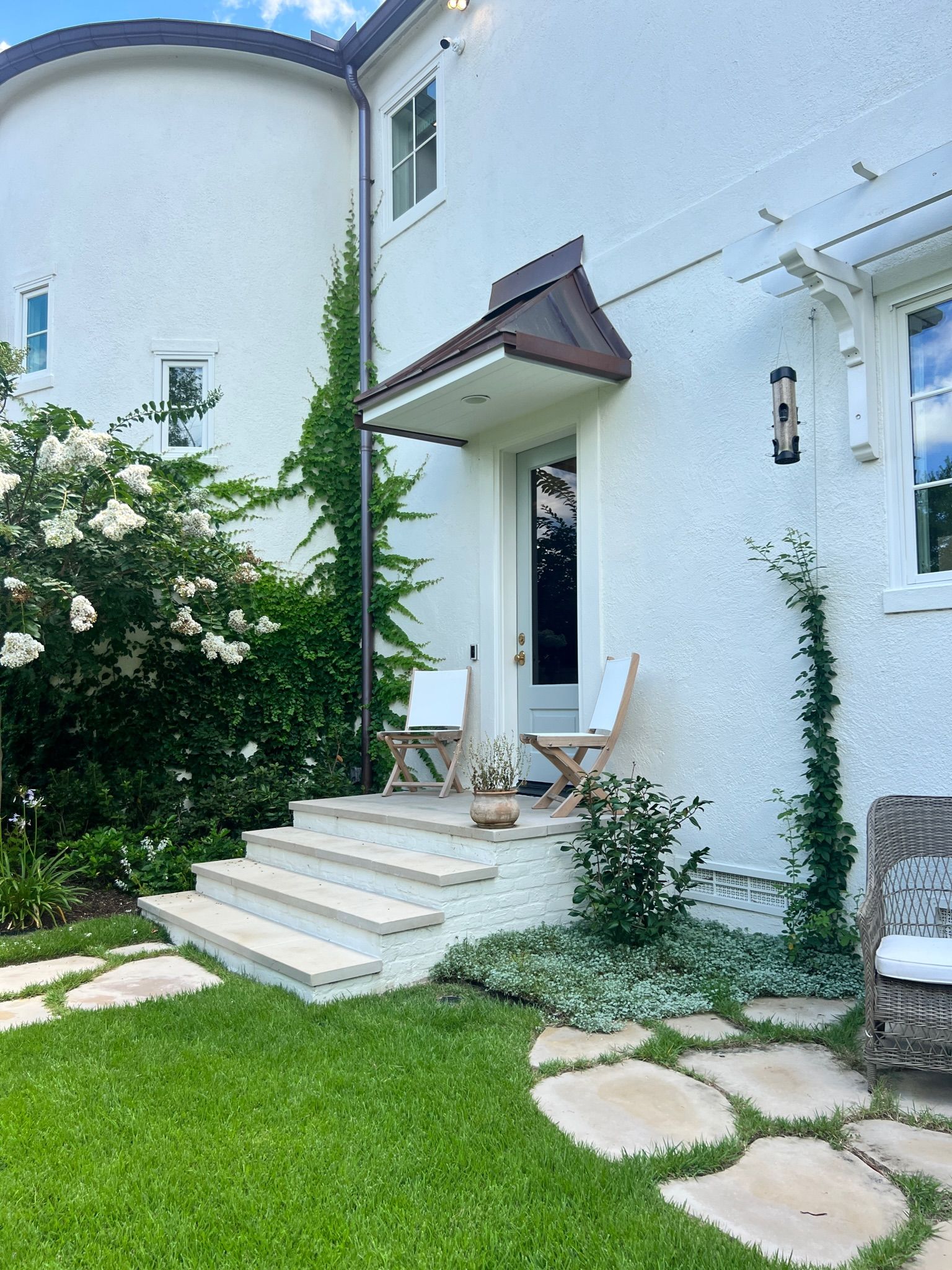 White stucco house entrance with steps, small awning, and green lawn with stepping stones