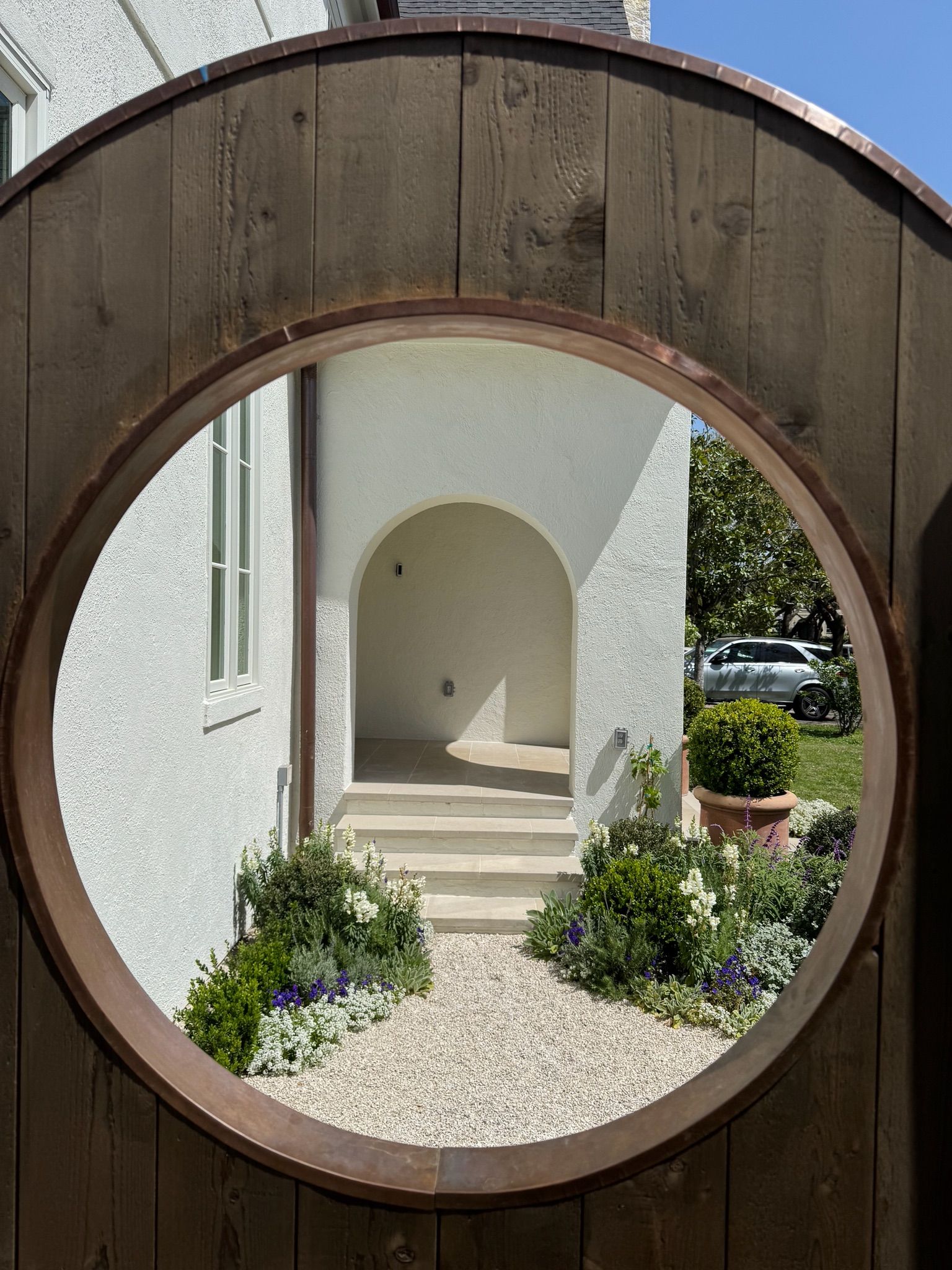 Circular wooden gate framing a white stucco courtyard with steps, archway, and flowering plants