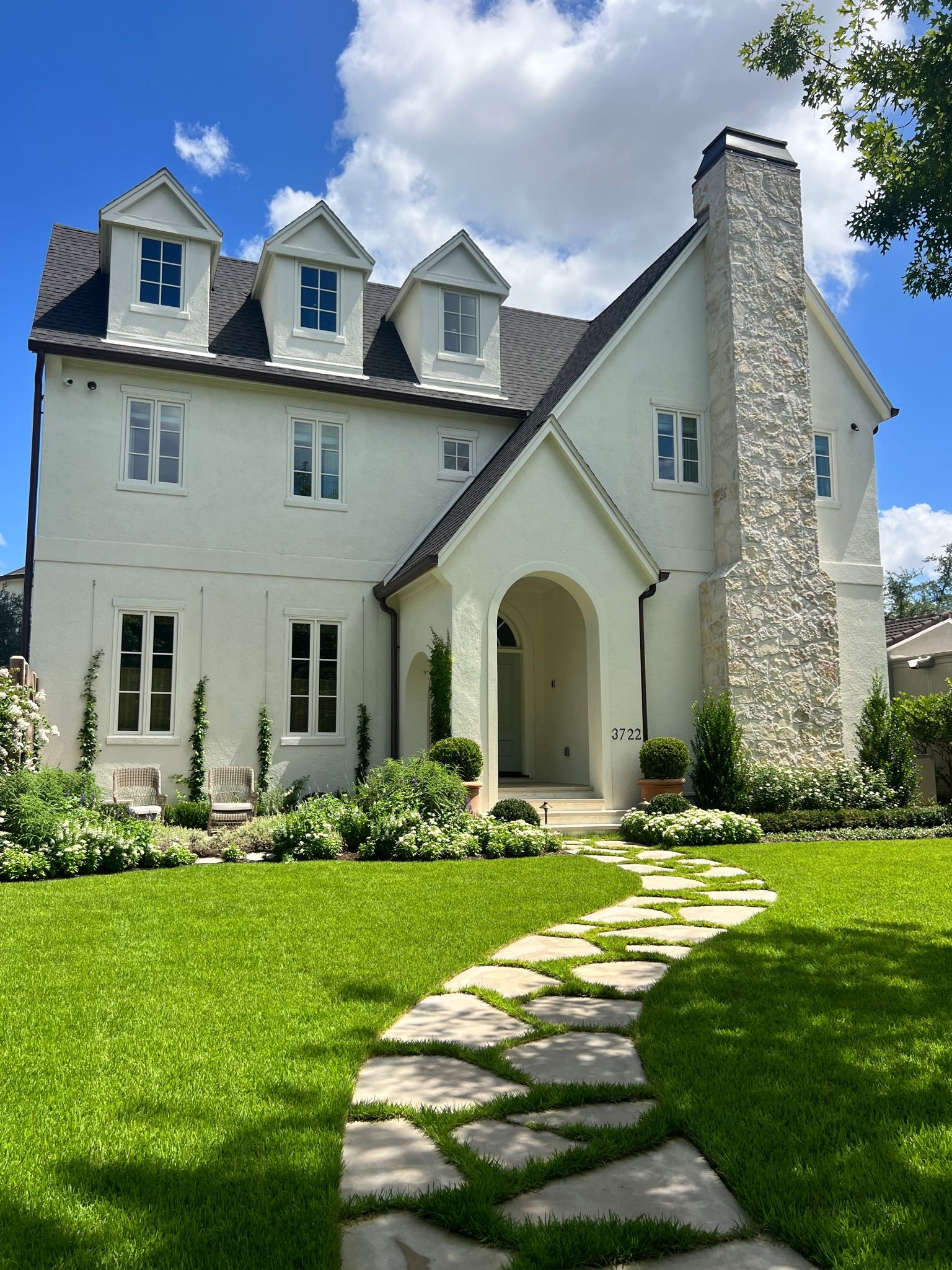 White stone house with arched entry, dormer windows, and a stone path through a green lawn under blue sky