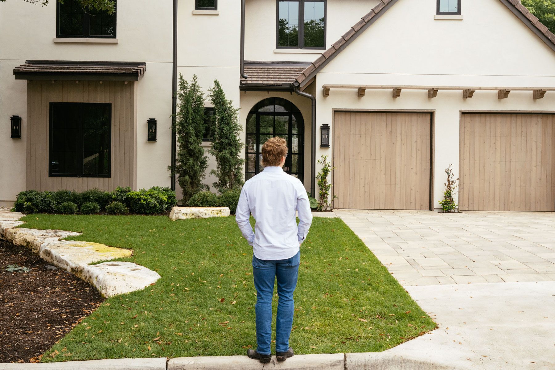 Person standing on a sidewalk facing a modern house with a lawn and driveway