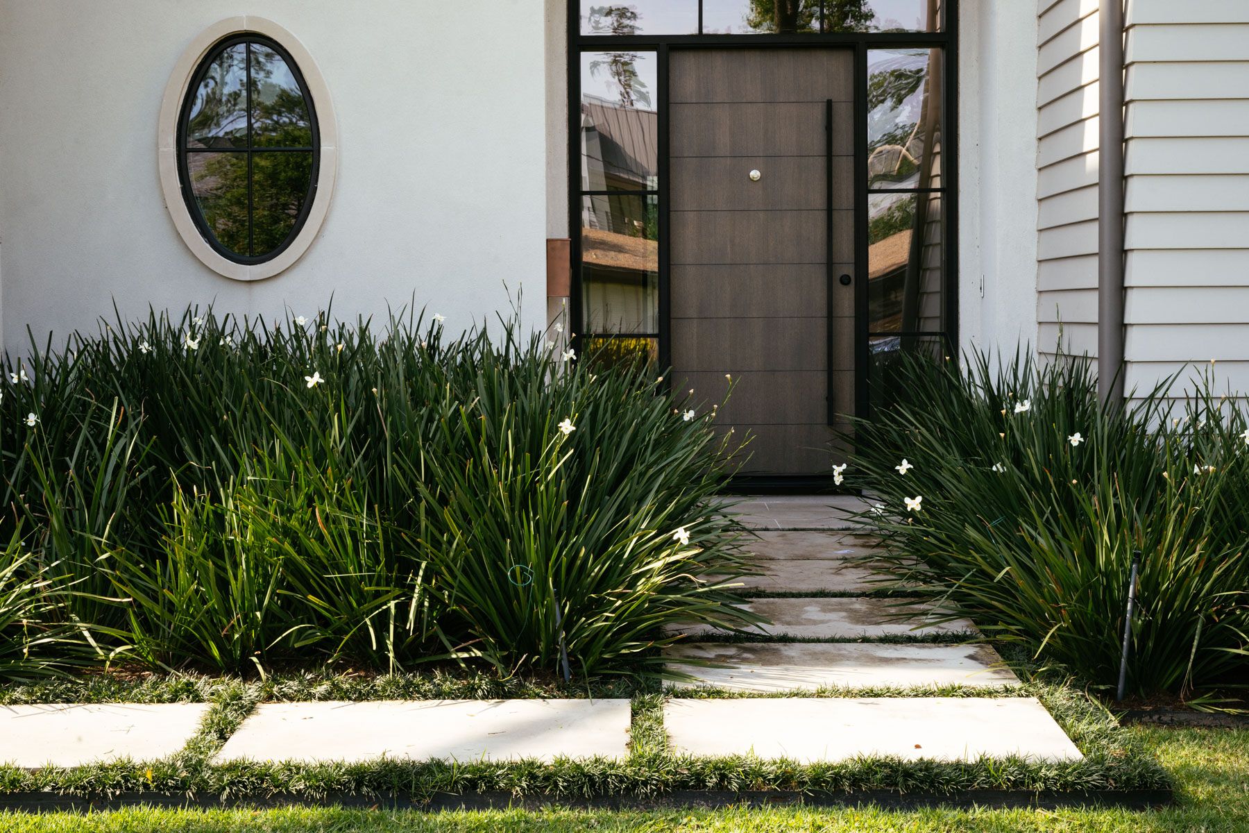 Front entrance with wooden door, oval window, and lush green landscaping along a stone path
