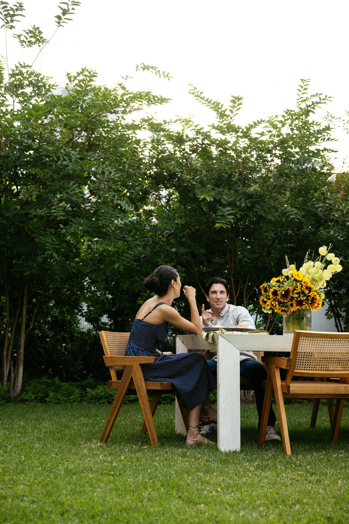 Two people sit at a white table on a grassy lawn, with chairs and sunflowers in a garden.