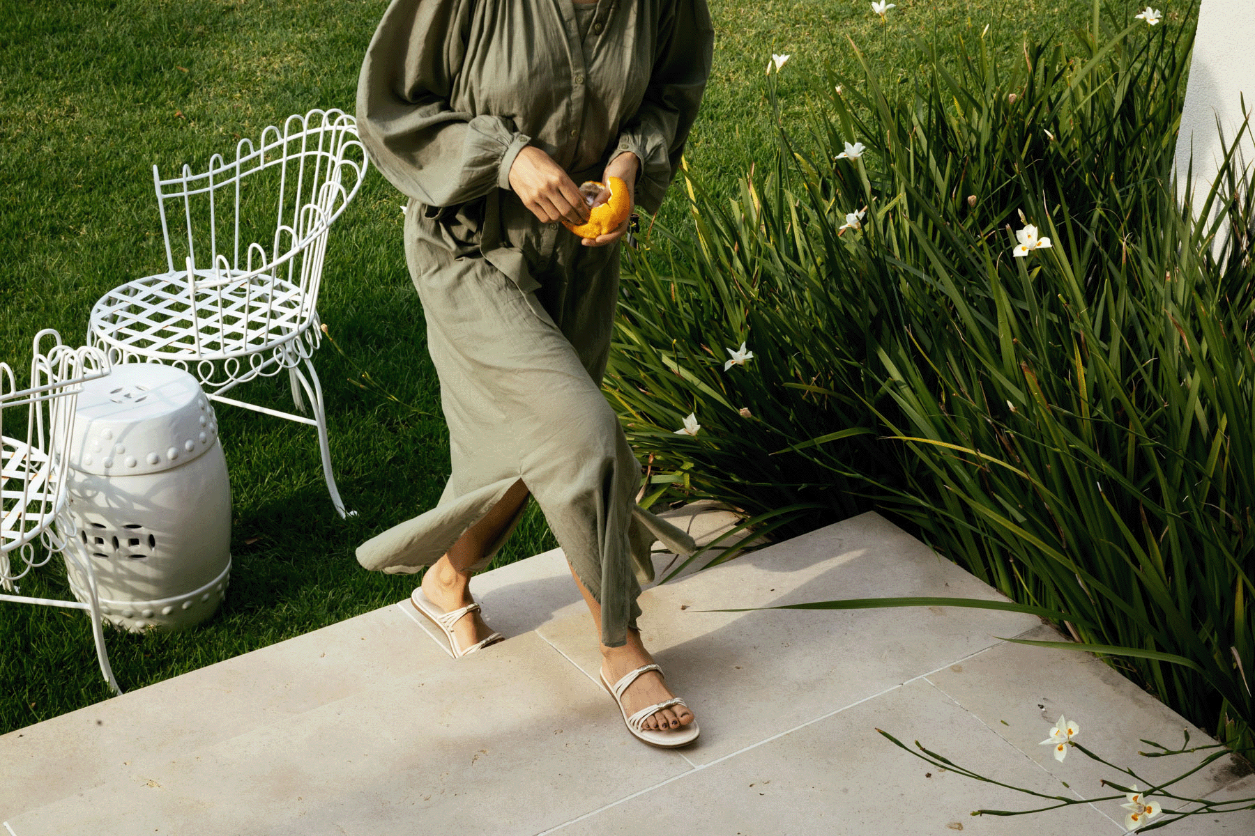 Person holding yellow flowers walks on a stone path beside a white chair and tall green plants.