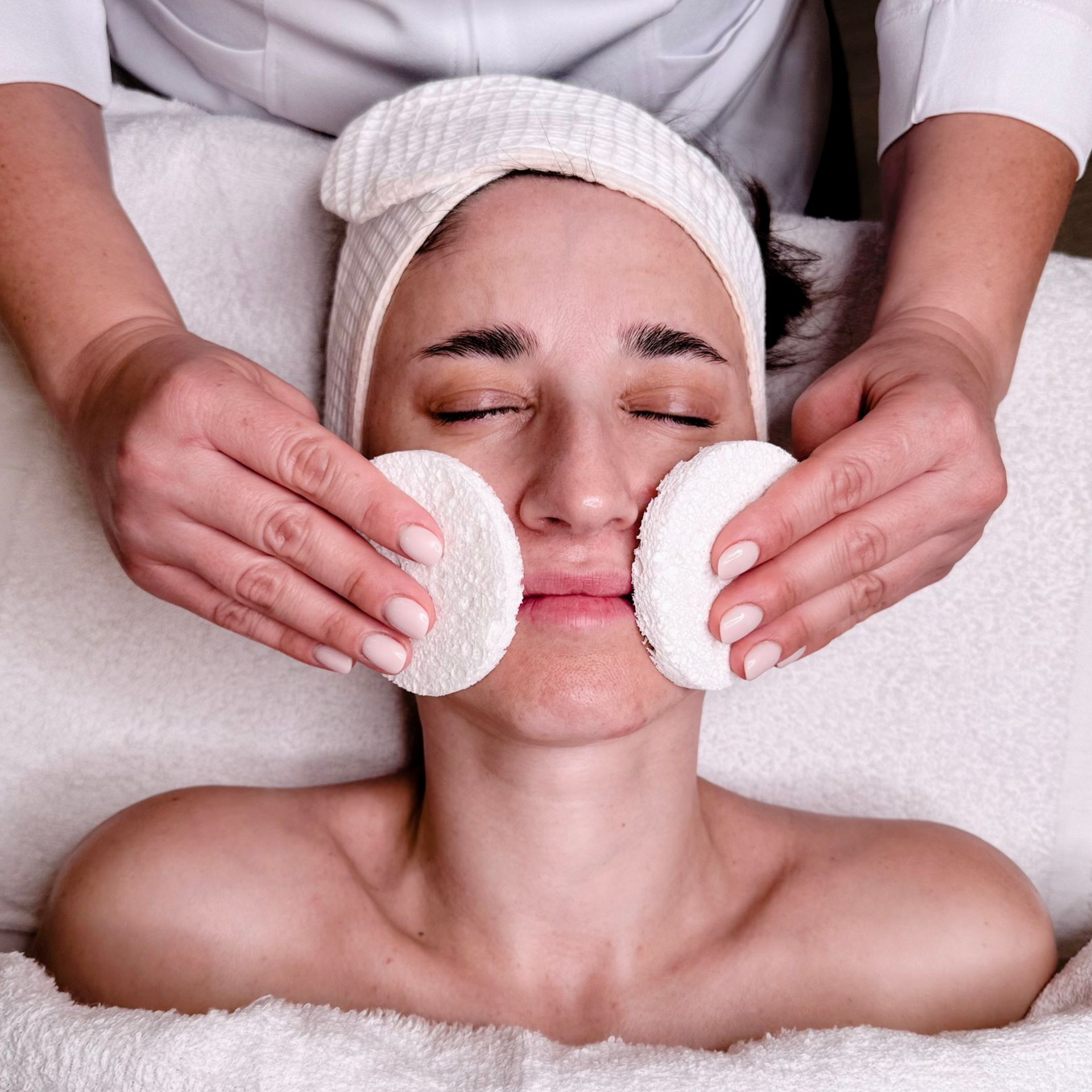 Woman receiving facial treatment at a spa, with eyes closed, cotton pads on her cheeks, and a therapist's hands holding them.