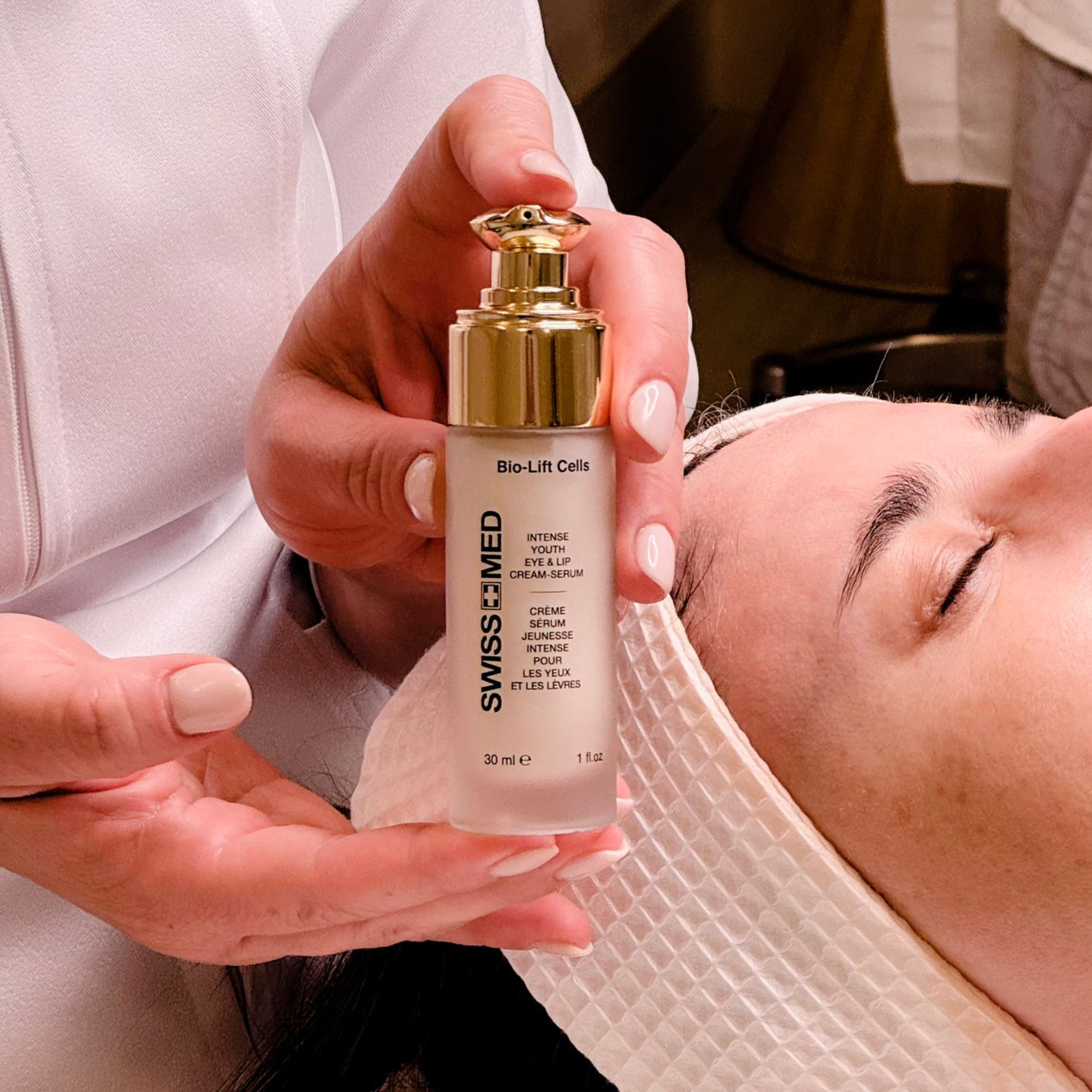 Woman holding Swiss+Med face serum bottle over a client's face in a spa setting.