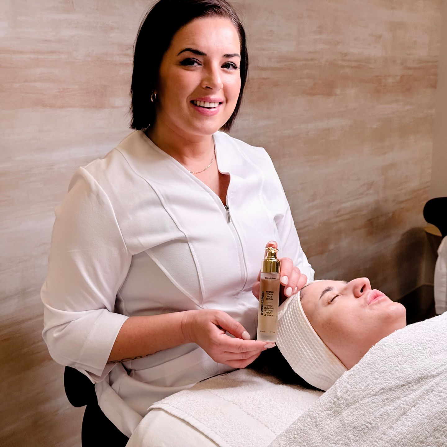 A woman in a white lab coat applies skincare to a client in a spa setting.