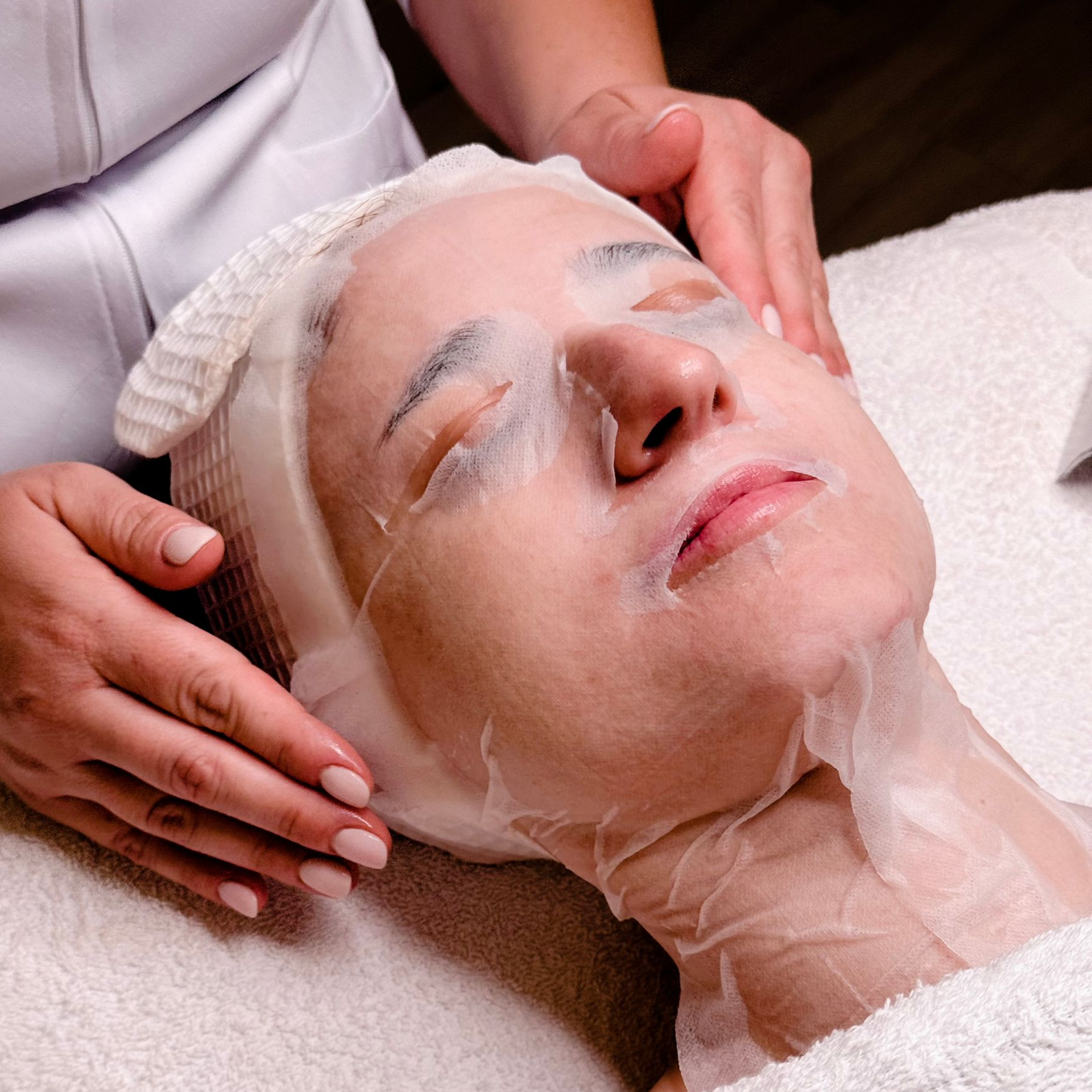 Woman receiving a facial treatment; sheet mask applied, hands gently massaging her face.
