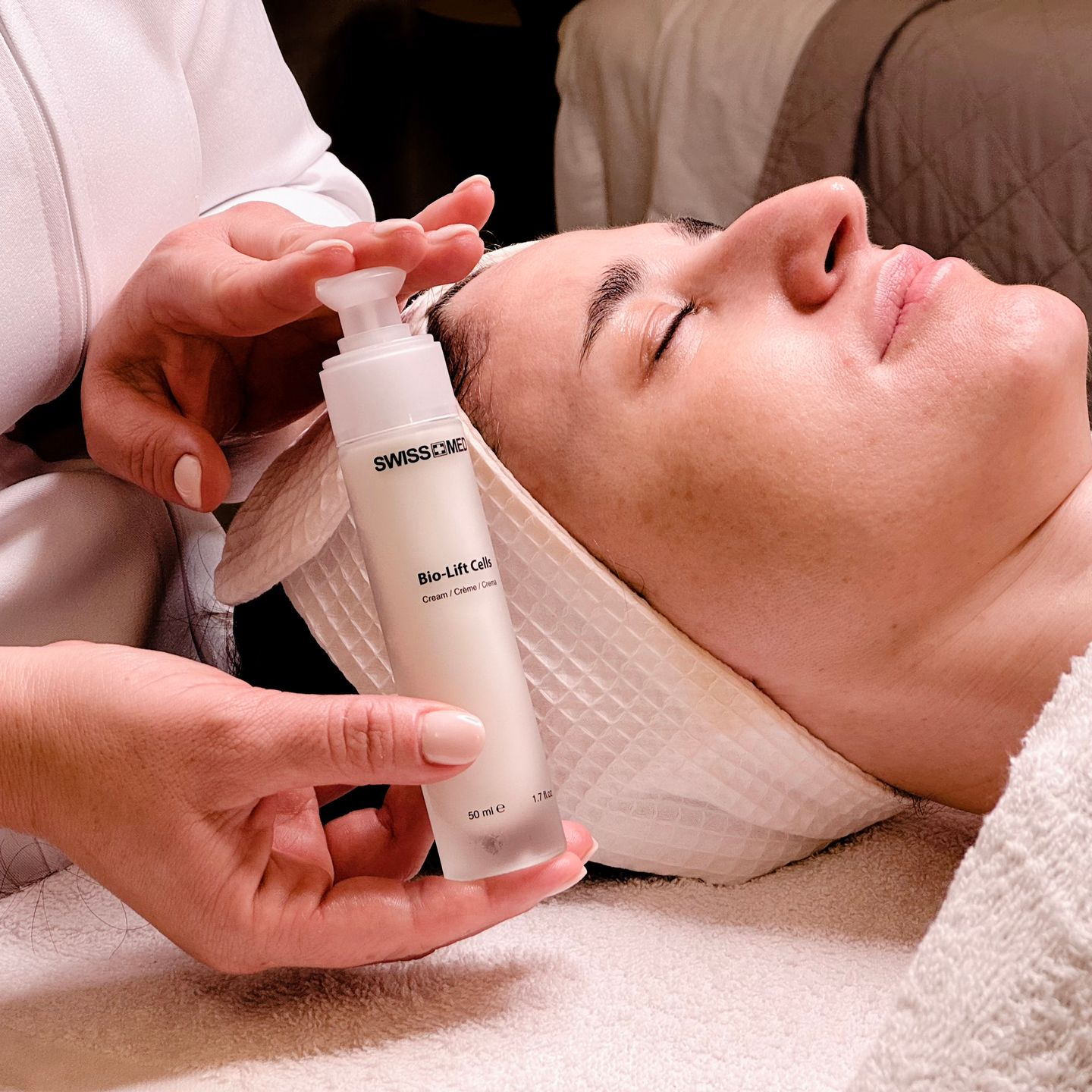 Woman receiving facial treatment with a bottle of moisturizer. The woman is lying down, eyes closed, at a spa.