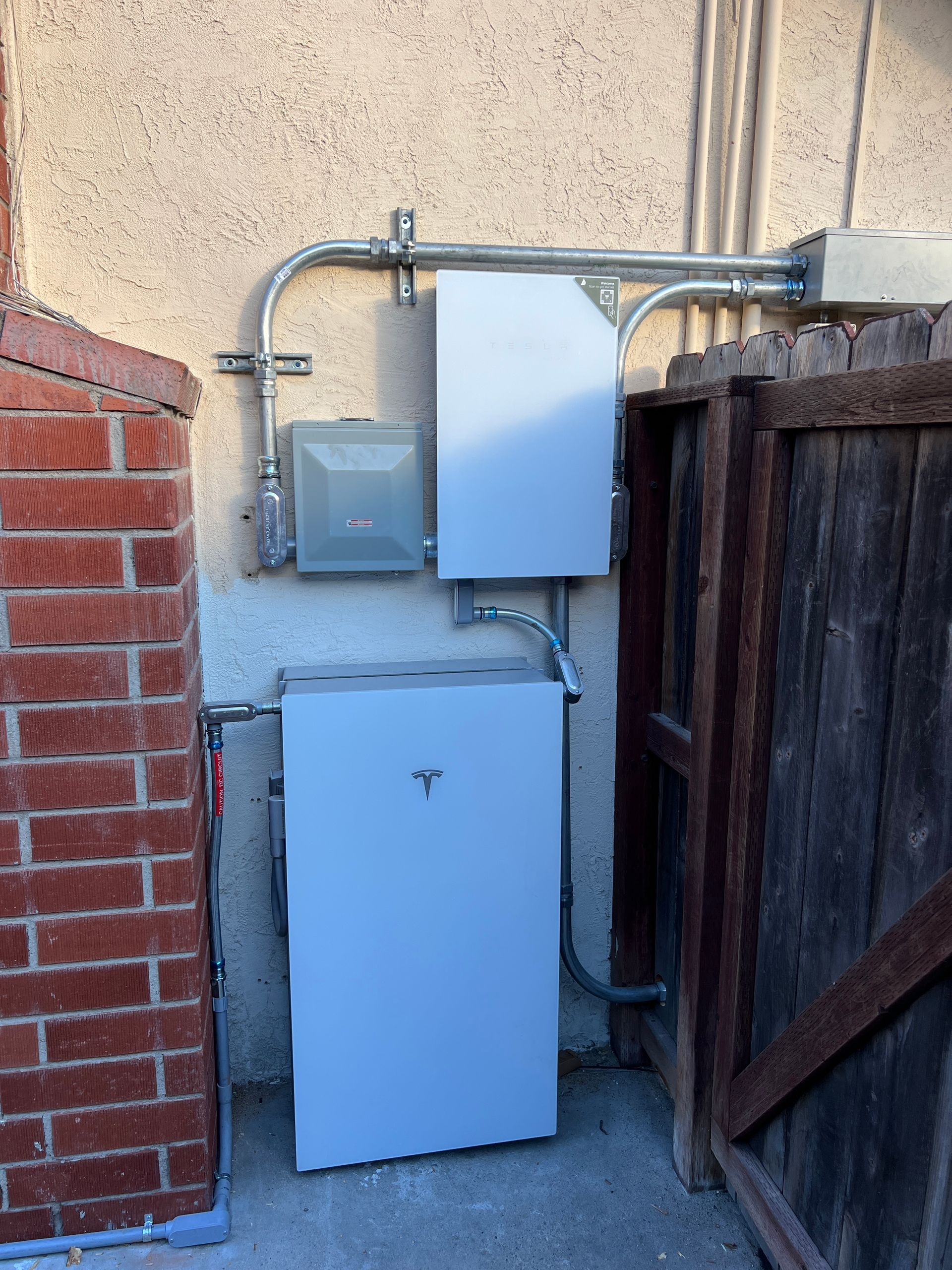 Two white utility boxes and a meter mounted on an exterior wall beside a wooden fence and brick wall.