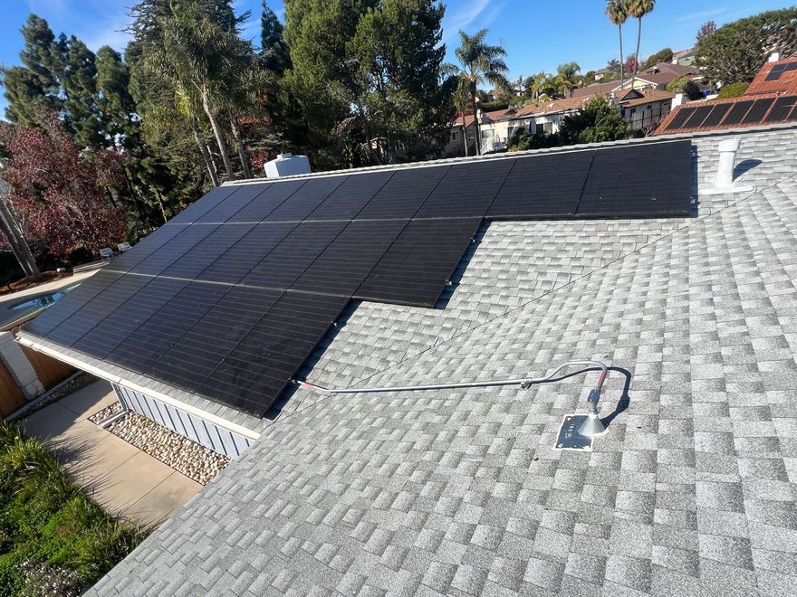 Solar panels on a sloped roof beside gray shingles, with a small tool on the roof under a clear sky.