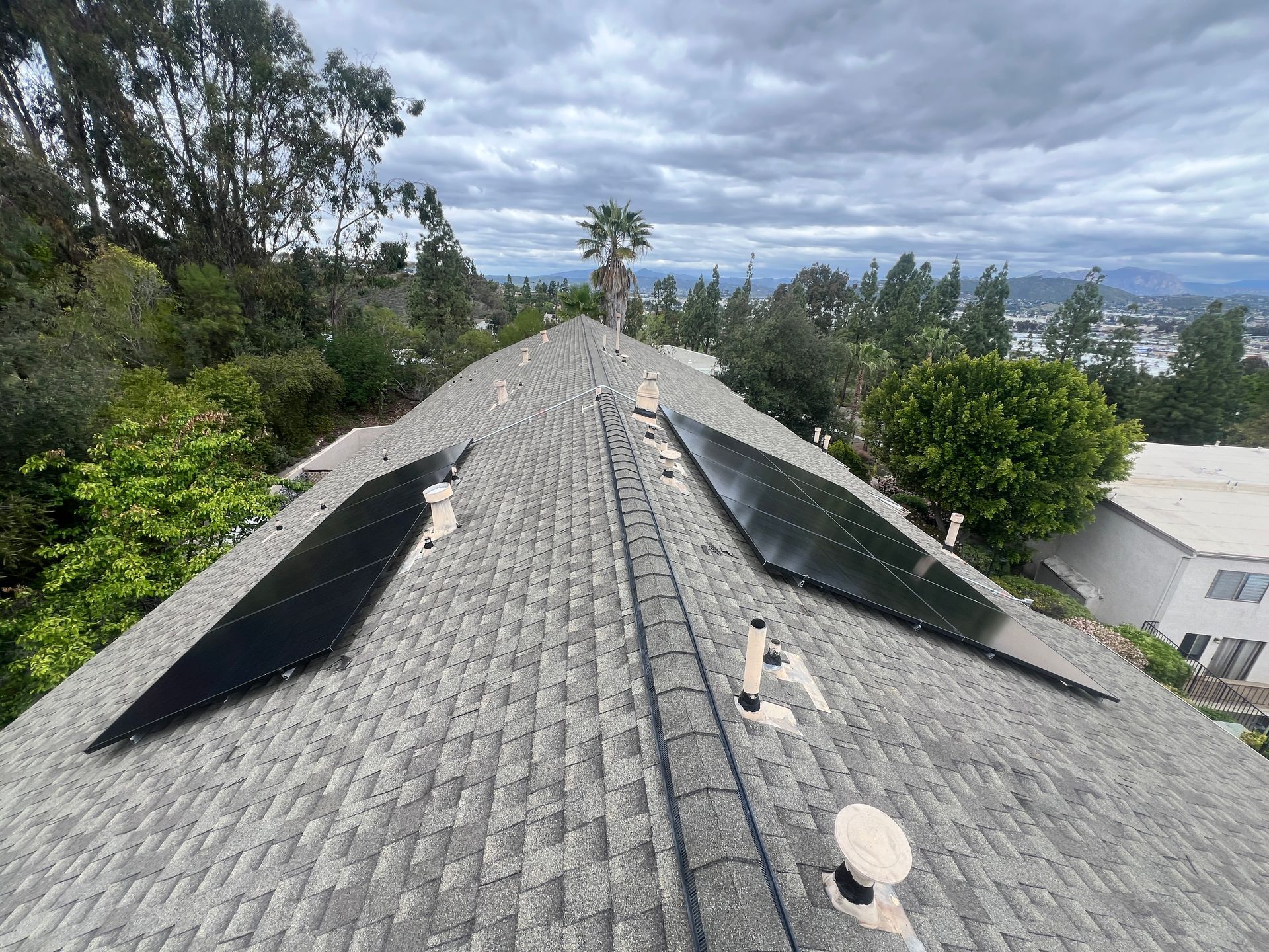 Roof with solar panels on a house, surrounded by trees and distant hills under a cloudy sky