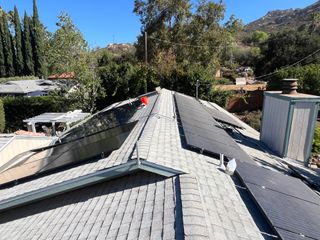Rooftop with gray shingles, chimneys, and a tree-lined hillside in the background