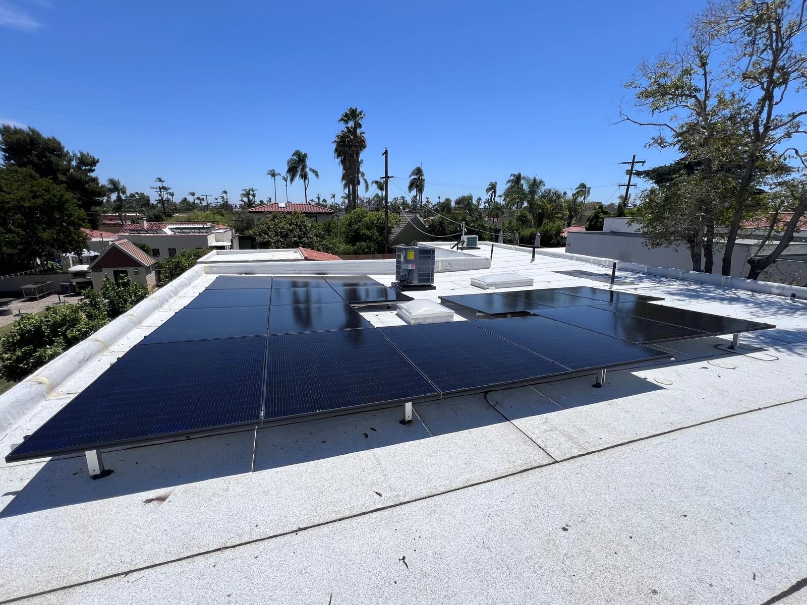Solar panels on a flat rooftop under a clear blue sky, with trees and houses in the background