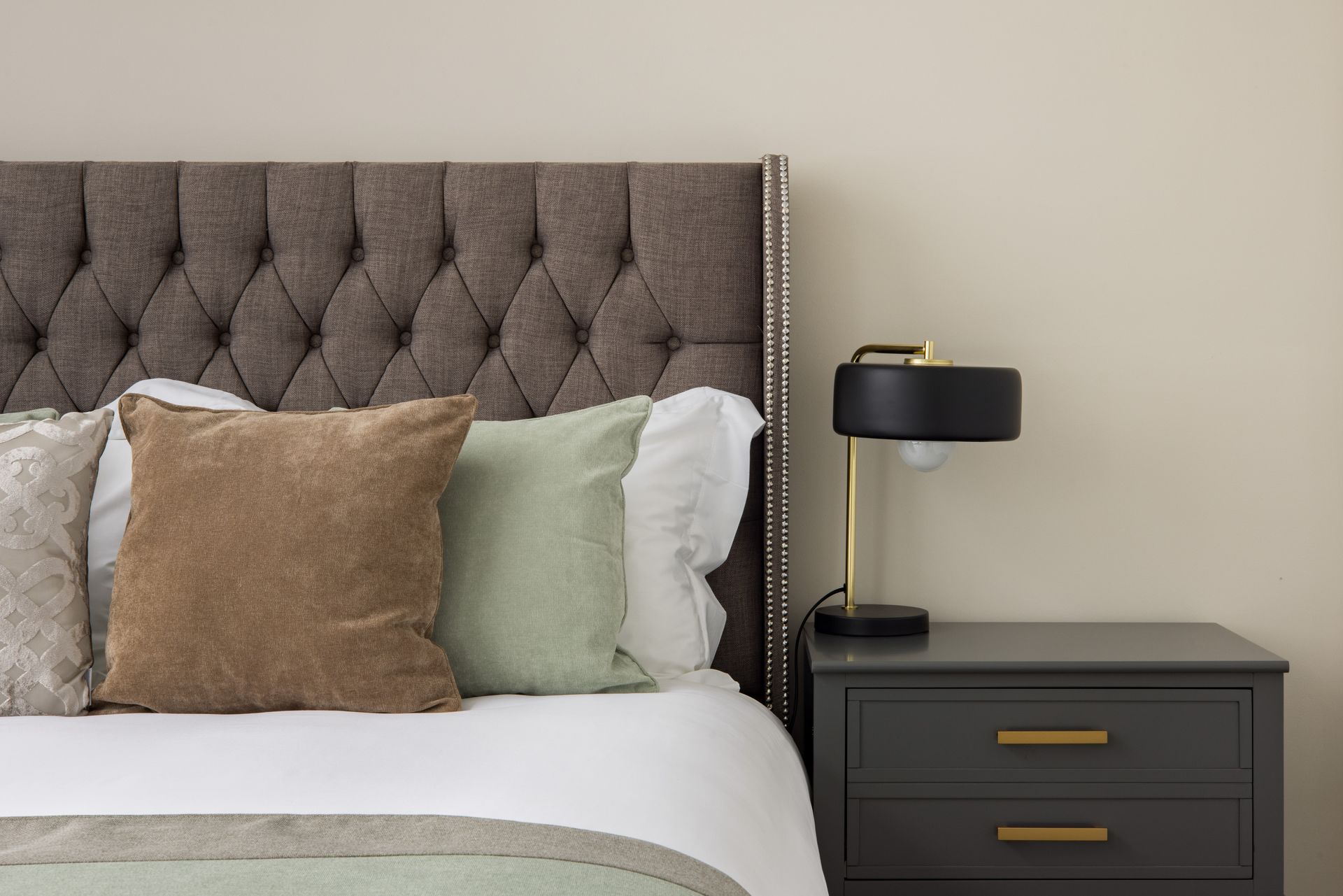 Bedroom with tufted beige headboard, white bedding, throw pillows, and a black nightstand with lamp.