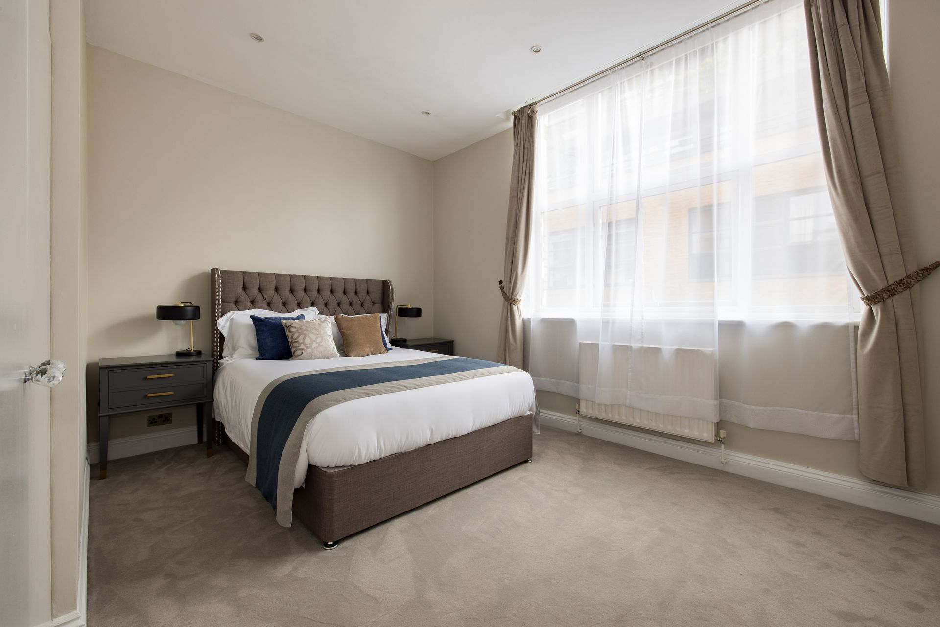 Bright bedroom with a neatly made bed, nightstands, and large sheer-curtained window