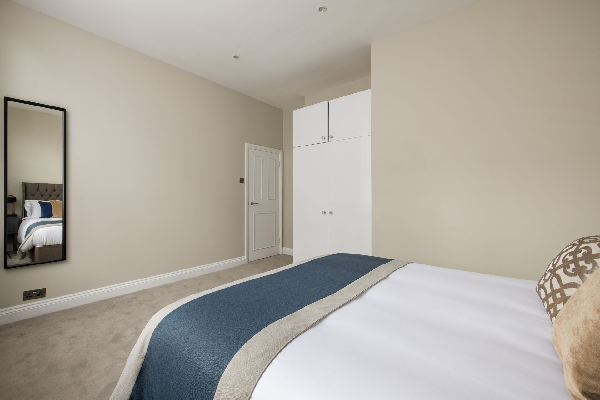 Minimal bedroom with beige walls, white doors, a wall mirror, and a bed with a blue-and-white blanket