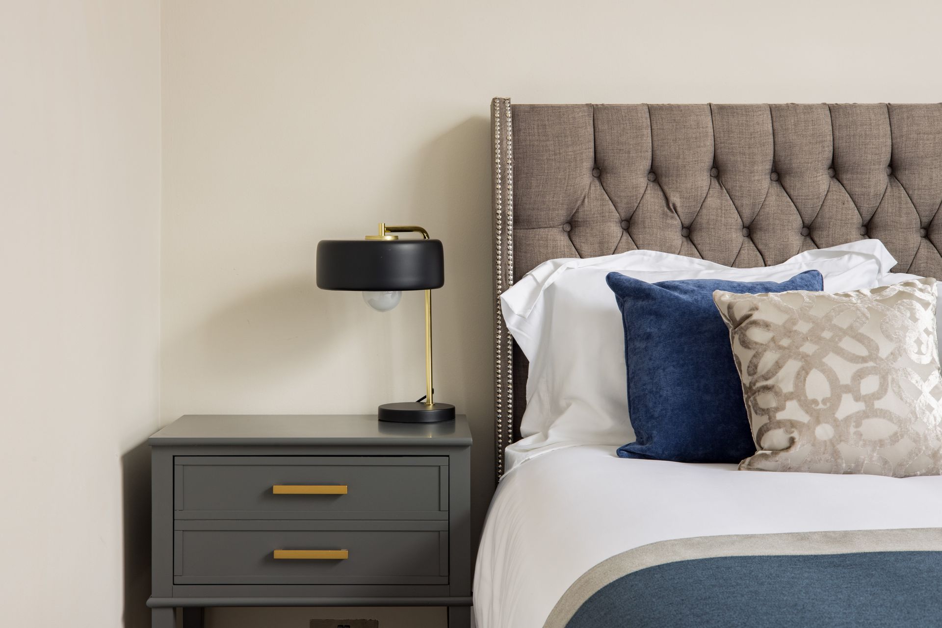 Modern bedroom with gray tufted headboard, navy lamp, and gray nightstand beside a bed with blue pillows