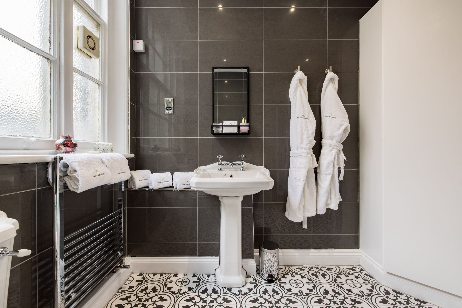 Modern bathroom with pedestal sink, dark tile shower wall, patterned floor, and white robes on hooks