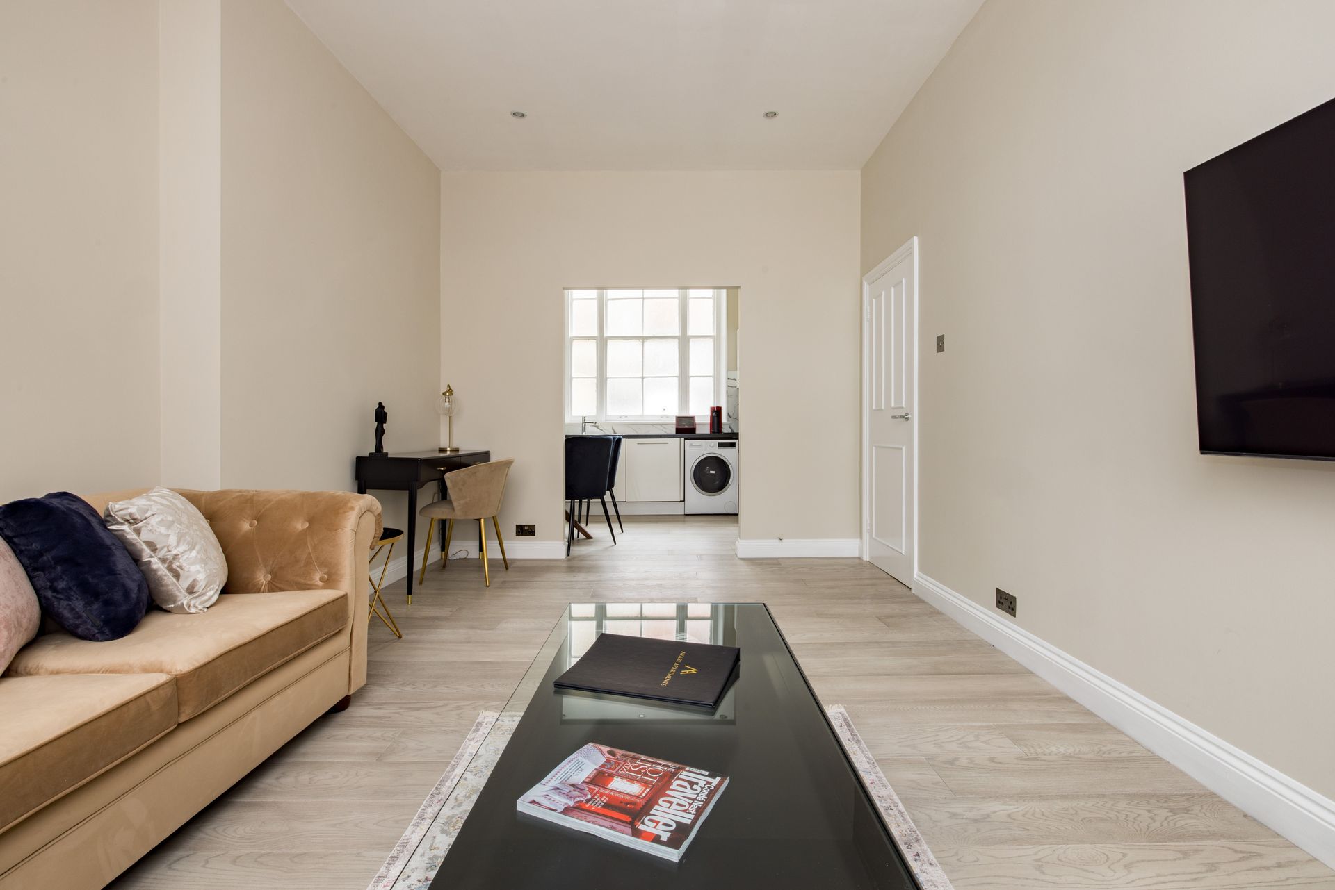 Bright living room with beige sofa, black coffee table, wall-mounted TV, and small workspace in the back
