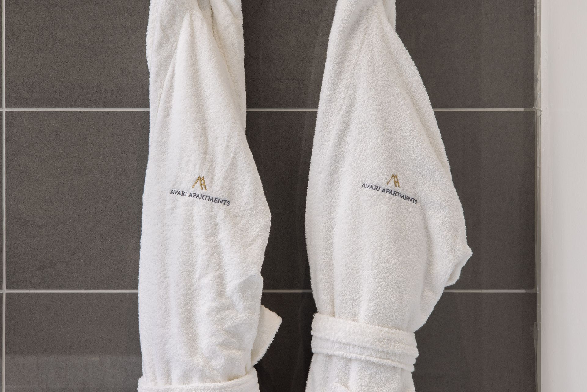 Two white bathrobes hanging side by side on dark tiled wall hooks