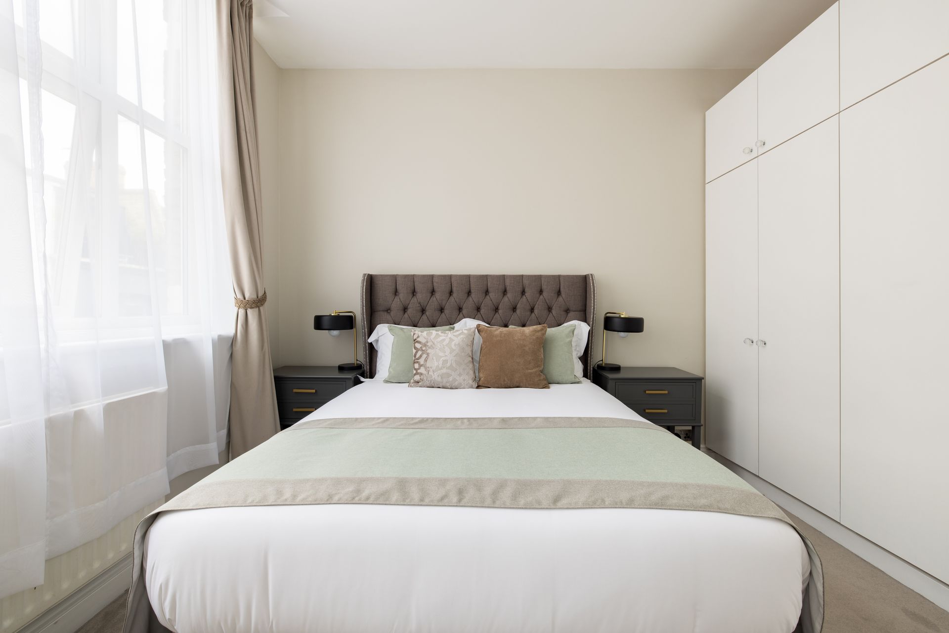 Bright bedroom with a tufted bed, white wardrobe, and large window with curtains.